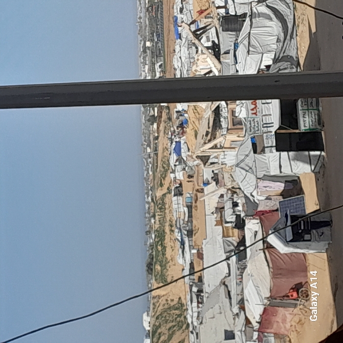 The image displays a densely populated informal settlement or refugee camp situated in Khan Yunis, Palestinian Territory, beneath a clear blue sky. The ground throughout is sandy dirt. In the foreground, numerous makeshift structures and tents, primarily white tarpaulin, are closely packed. One individual, dressed in dark clothing with white stripes, stands near a white tent where a blue solar panel leans against it. To the immediate right, another white tent has clothes drying on its exterior. Further into the mid-ground, a red two-wheeled cart is visible near a tent. Multiple structures appear partially collapsed or damaged, revealing exposed wooden supports. A white tent in the mid-right features signs with Arabic text, including "لصرافة و التحويلات المالية" (For money exchange and financial transfers) and "PYT PAY." A small cluster of three to four individuals is observed walking on a dirt path in the mid-distance. The upper left quadrant of the frame is crossed by black electrical wires, and a thick, dark vertical pole partially obstructs the right view. The background shows an undulating sandy landscape dotted with more tents and distant structures extending to the horizon.