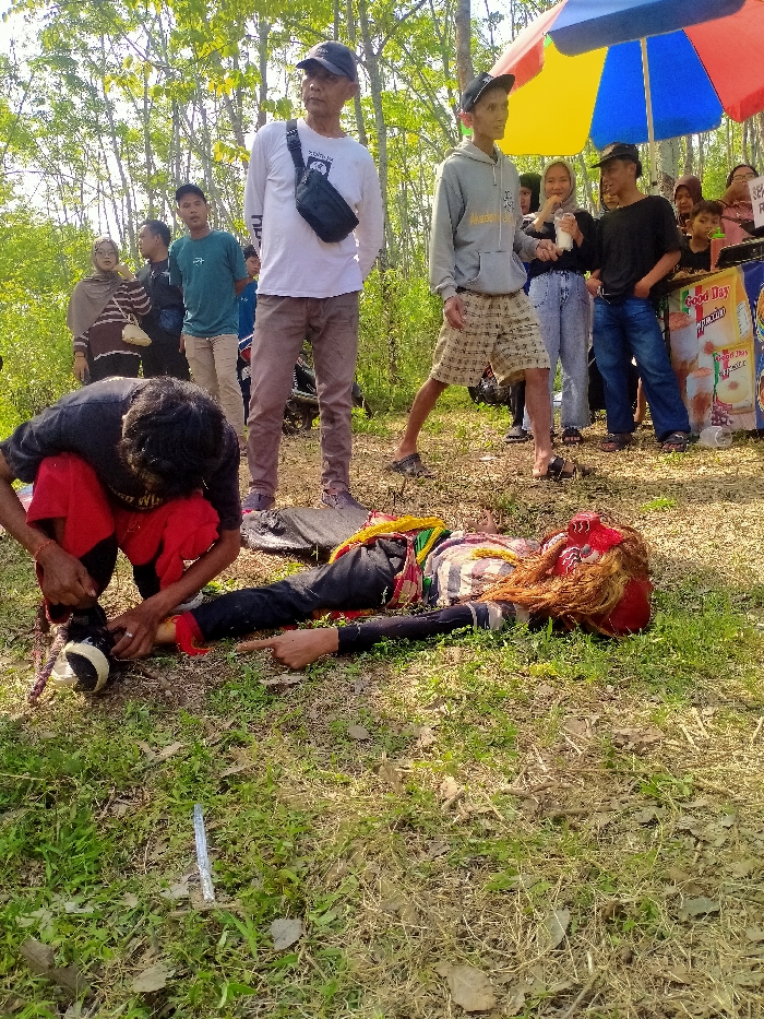 The image captures an outdoor scene in Tawangsari, Indonesia, on a sunny day with patchy dry grass covering the ground and sparse trees with light green foliage in the background.
In the foreground, a person lies supine on the grass, wearing dark pants, a striped long-sleeve top, and an elaborate red mask adorned with long reddish-brown hair. Yellow and green fabric strips are draped around the upper torso. The individual's left arm is extended forward with the index finger pointing. To the left, a man in a black t-shirt and red shorts kneels, actively adjusting or securing a black and white sneaker on the prone person's left foot. A long, thin, reflective metallic object rests on the grass near the outstretched hand of the prone individual.
In the mid-ground, several individuals are observing the scene. A man wearing a white long-sleeve shirt, dark pants, and a black baseball cap stands, looking towards the right, with a black fanny pack slung across his chest. To his right, a slender man in a grey hoodie, patterned shorts, and dark sandals stands, looking left. Further to the right, a vendor stall is visible, topped with a multi-colored (red, yellow, blue) umbrella. The stall features promotional graphics, including the "Good Day" brand. Several people are gathered near the stall; one person is holding a clear cup containing a white beverage with a straw, and a man in a black t-shirt and blue jeans stands next to the stall. Additional onlookers are partially visible in the left background, including a woman in a brown hijab and striped top. A motorcycle is partially visible behind the man in the white shirt.