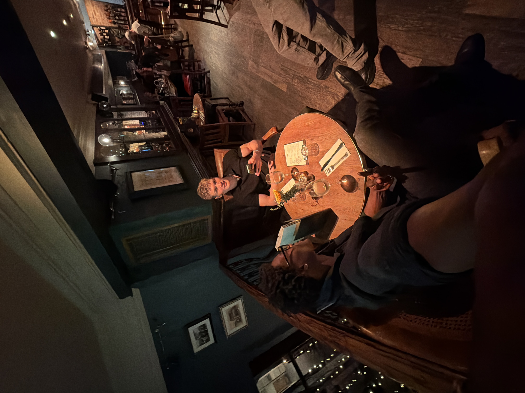 The image captures an indoor evening scene in a dimly lit, cozy establishment, likely a bar or restaurant in London. The ambiance is warm and inviting, with dark walls, wooden furniture, and soft lighting.
In the foreground, two individuals are seated at a round wooden table. To the left, a person with light, curly hair, wearing a dark shirt, looks towards the camera with a relaxed expression, holding a drink. Opposite them, another person with dark, curly hair and glasses is looking down, seemingly at a tablet or a menu held open in their hands. On the table between them are several wine glasses, a small vase with yellow flowers, cutlery, and a menu with the word "DRINKS" clearly visible at the top. The legs and feet of other people are visible, suggesting more individuals are seated around the table or nearby.
The background reveals a bustling interior with more patrons seated at various tables. Shelves along the walls are adorned with bottles, glassware, and other decorative items, including what appears to be a bell jar or similar scientific instrument. Framed pictures hang on the dark walls, and a ventilation grate is visible above the heads of the foreground subjects. String lights or fairy lights add a festive, intimate glow to the lower right portion of the image. The overall impression is one of a social gathering in a comfortably upscale setting during the evening.