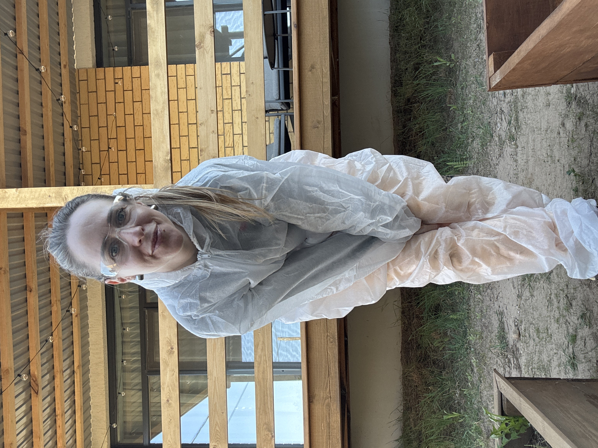 A person in a full-body white protective suit, wearing safety glasses, is kneeling on a dirt and grass surface adjacent to a structure with wooden beams and brickwork. The person is looking at the camera and smiling. The structure appears to be a building or part of a building with windows. There are string lights along the wooden beams. The photograph was taken in the city of Chaikovsky, Russia.