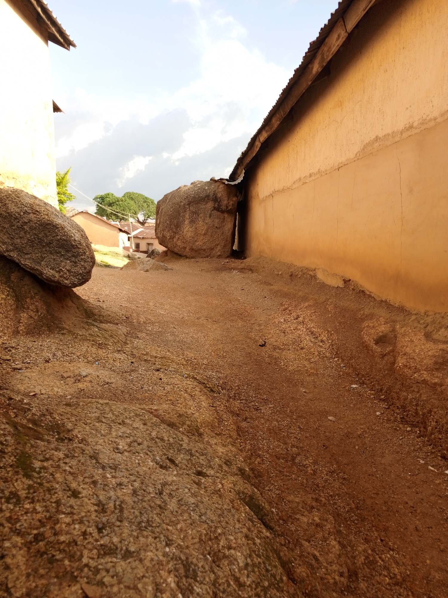 A low-angle, wide shot captures a narrow, dirt path between a large boulder on the left and the ochre-colored wall of a building on the right. The path is covered in gravel and small stones, with a few scattered leaves. The boulder on the left is rough and textured, with mossy patches. Further back, a large, rounded boulder is nestled against the building's wall, creating a dark, narrow opening between them. The building's wall is rough and cracked, with a corrugated metal roofline visible at the top. In the distance, beyond the boulders, several small, tan-colored buildings with similar corrugated roofs are visible, interspersed with lush green trees. The sky above is a pale blue, with large, fluffy white clouds. The overall impression is of a dry, dusty, and rustic environment.