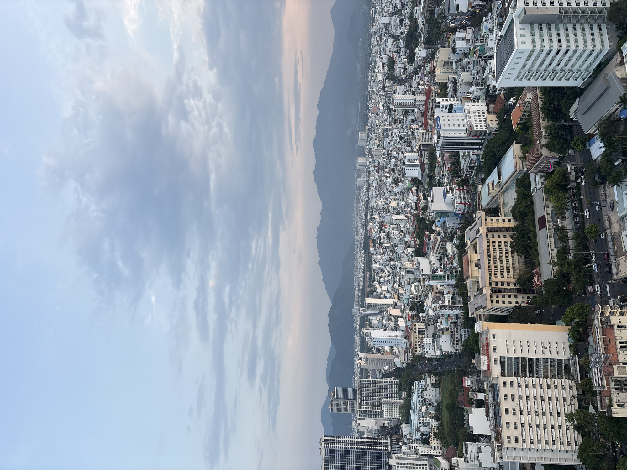 The image depicts a high-angle view of a densely populated urban area adjacent to a mountain range. The city, presumably Nha Trang Ward, Vietnam, is composed of numerous buildings, varying in height from low-rise structures to skyscrapers. The buildings are primarily white or light-colored. The sky is filled with clouds.