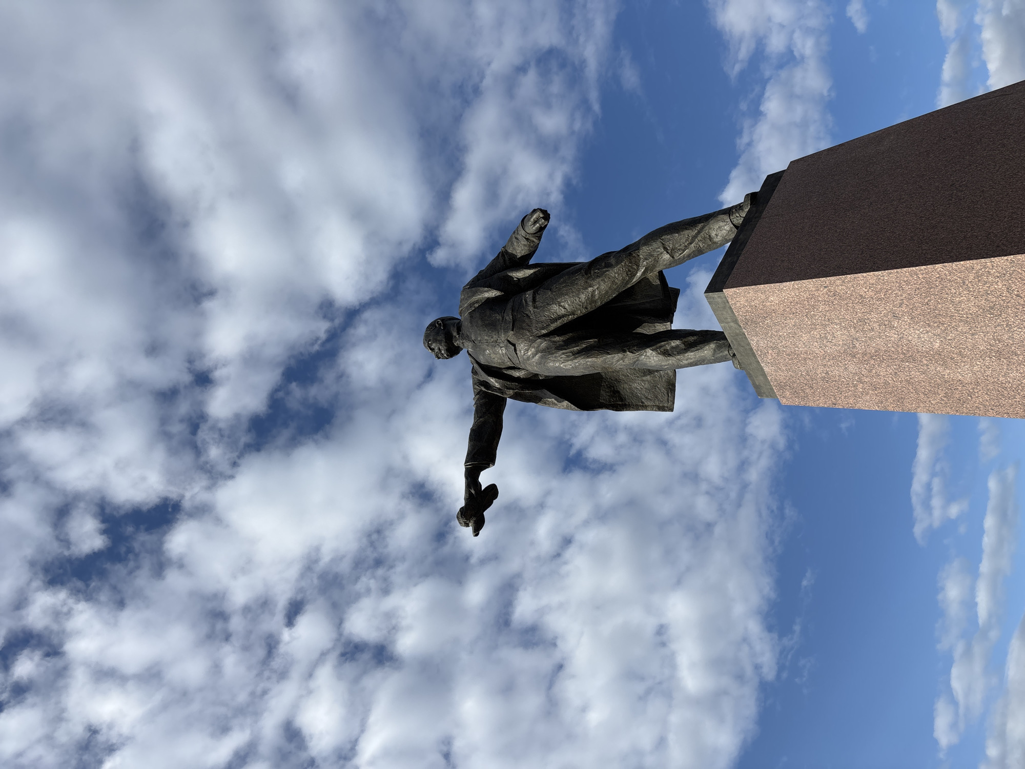 The image depicts a bronze statue of Vladimir Lenin in Saint Petersburg, Russia. The statue stands atop a large, rectangular pedestal made of red granite. Lenin is portrayed in a long coat, with one arm raised and a cap in the other hand. The perspective is from below, looking up at the statue, with a blue sky filled with scattered white clouds in the background.