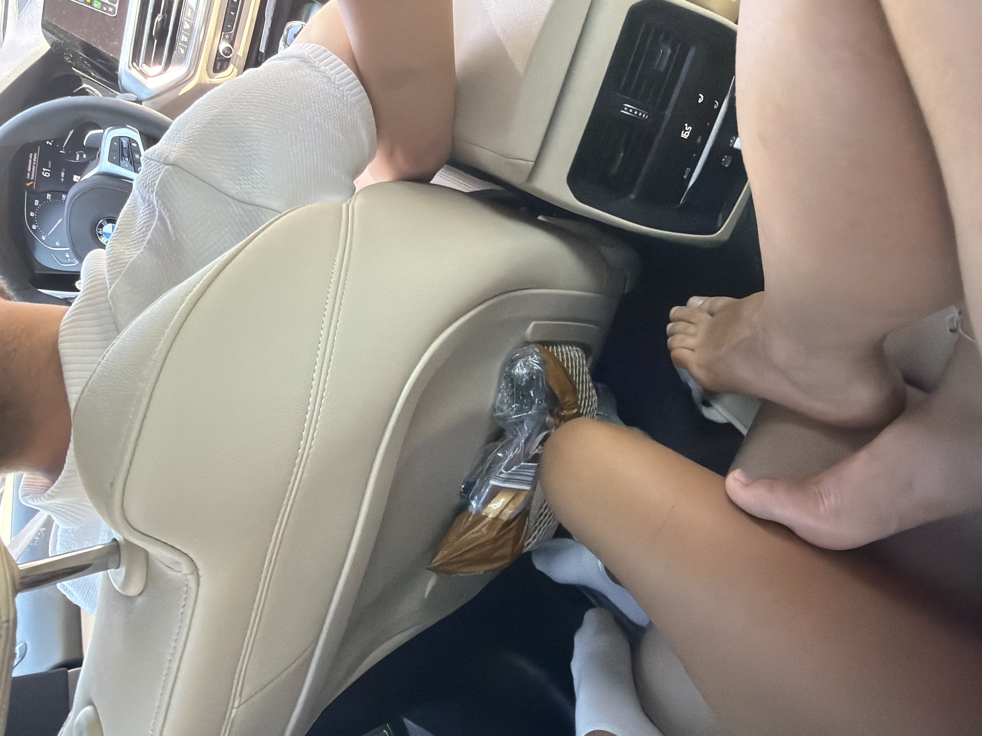 The image shows the interior of a car from a low angle, focusing on the legs and feet of passengers in the back seat. The car's interior is upholstered in light-colored leather. A person in the front seat, wearing a white shirt, is visible from behind, with their arm resting on the back of the front seat. In the back seat pocket, there's a package of food, possibly snacks. Two sets of feet are visible in the foreground, one with white socks and the other bare. The car's rear climate control panel is visible, showing a temperature of 16.5 degrees Celsius. The scene takes place inside a vehicle, but the specific location within the city of Târgu Secuiesc, Romania, cannot be determined from the visual information provided.