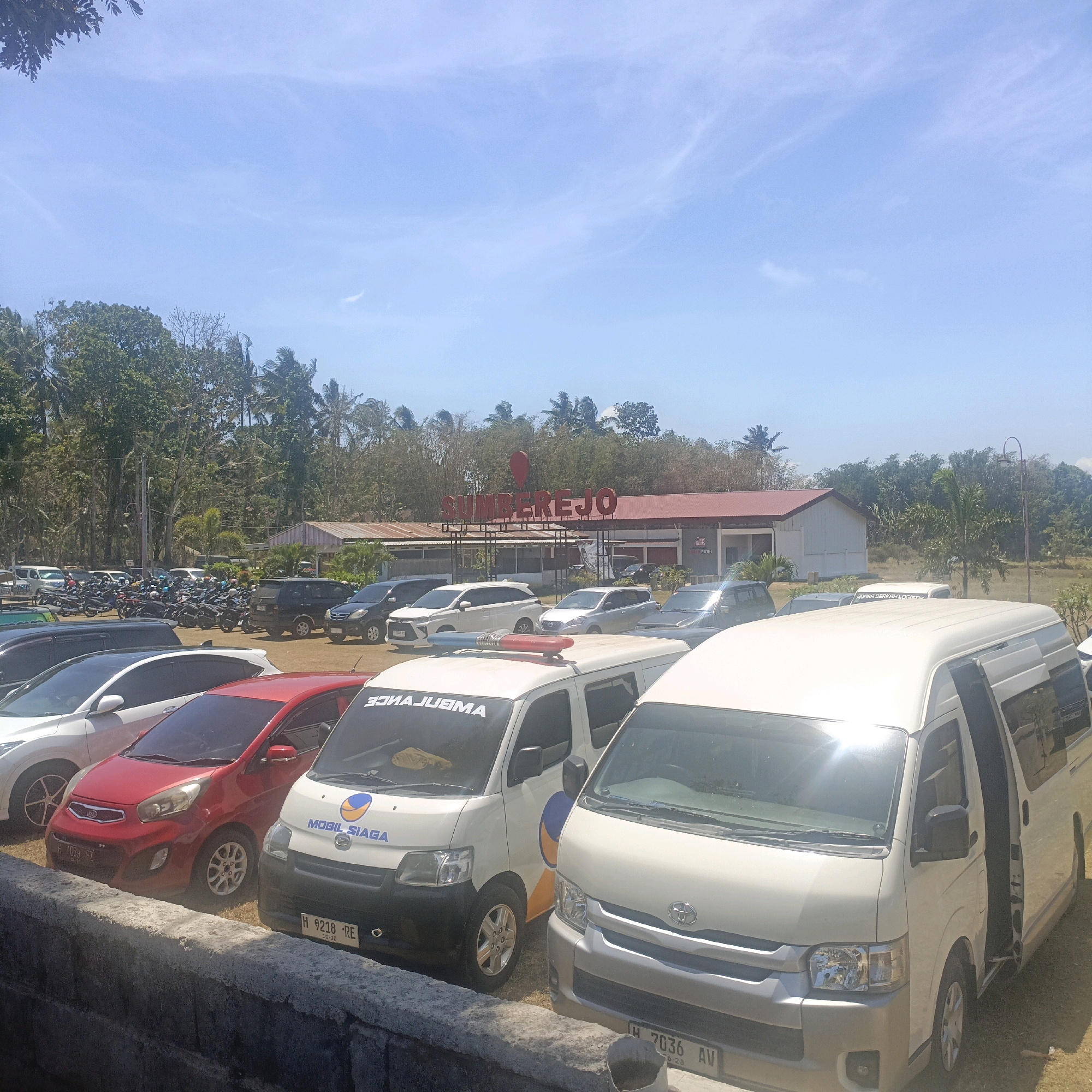 The image captures a sun-drenched outdoor scene in Krajan Lor, Indonesia. In the foreground, a low concrete wall separates a grassy area from a parking lot. Parked vehicles, including a red Kia hatchback, a white hatchback, and two white vans, are visible. One of the vans is an ambulance with its hazard lights flashing, a blue and red light bar positioned above the windshield. The text "AMBULANCE" is visible on the windshield in reverse, and "MOBIL SIAGA" is written on the side. The other white van has its passenger door open.
In the middle ground, a paved parking area is filled with motorcycles and several cars, including black SUVs and white sedans. Beyond the parking lot, a building with a red roof and white walls stands. A prominent sign with red block letters spelling "SUMBEREJO" is mounted on a metal structure in front of the building. Palm trees and other lush green foliage surround the building and extend into the background. The sky above is a clear, pale blue with scattered wispy clouds. The overall impression is that of a busy parking area for a facility, likely a public venue or business, in a tropical environment.