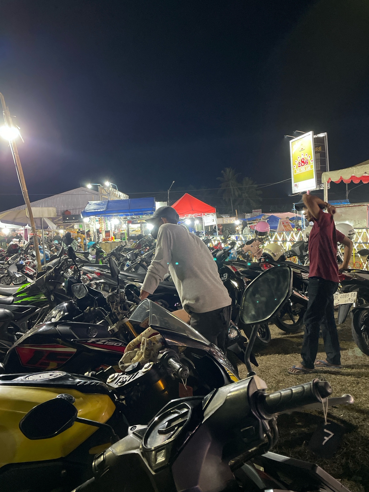 This is a nighttime, eye-level, medium shot of a crowded motorcycle parking area. The scene appears to be in Daya Asri, Indonesia, possibly at a night market or event, given the presence of food stalls and signage. In the foreground, several motorcycles are parked closely together, with their handlebars and front sections visible. The ground is covered in dry grass and dirt.
Two men are the primary subjects. One man, on the left side of the frame, is facing away from the camera, wearing a light-colored long-sleeved shirt and dark pants. He appears to be interacting with the motorcycles. Another man, on the right, is facing left and slightly away from the camera, wearing a maroon t-shirt and dark pants. He is raising his right arm, possibly stretching or gesturing. He is wearing flip-flops.
In the background, there are numerous other motorcycles, creating a dense parking lot. Beyond the motorcycles, there are various stalls and structures, some covered by colorful tents (red and blue are visible). Bright lights illuminate the area, suggesting commercial activity. A large vertical billboard with colorful graphics and Indonesian text is visible in the upper right quadrant. Palm trees are faintly visible in the distance against the dark night sky. The overall atmosphere is busy and illuminated.