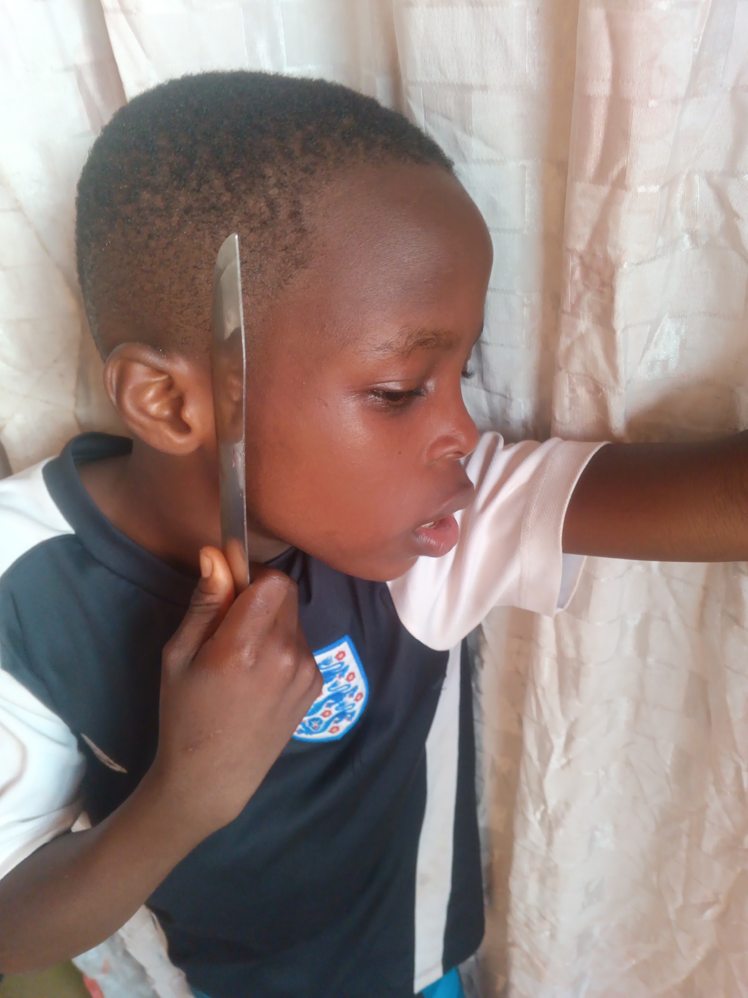 A young child with dark skin and short hair is depicted in a profile view against a light-colored, textured fabric background. The child is wearing a dark-colored short-sleeved shirt with white accents and a crest on the left chest. The child's right hand grasps the handle of a silver-colored metallic knife, positioning the blade vertically alongside the left side of their face, adjacent to the ear and cheek. The child's mouth is slightly open, and their gaze is directed downwards and to the right. The background appears to be an indoor curtain or draped textile. No specific visual elements confirm the city of Onitsha, Nigeria.
