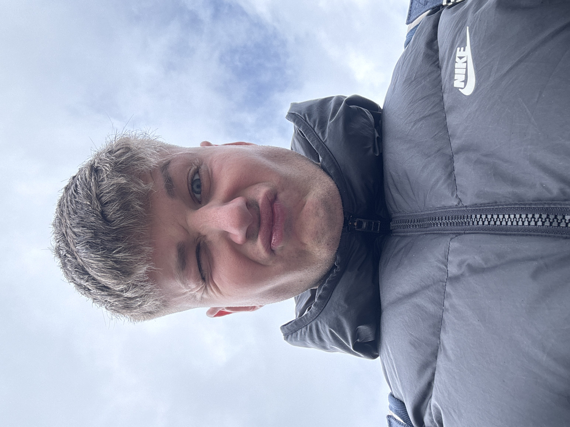 A close-up shot, taken from a low angle, depicts a man with short, blonde hair wearing a dark gray Nike puffer jacket. He is winking with his left eye, and his expression is a slight grimace or pout. The background is a cloudy sky. The location cannot be determined from the image.