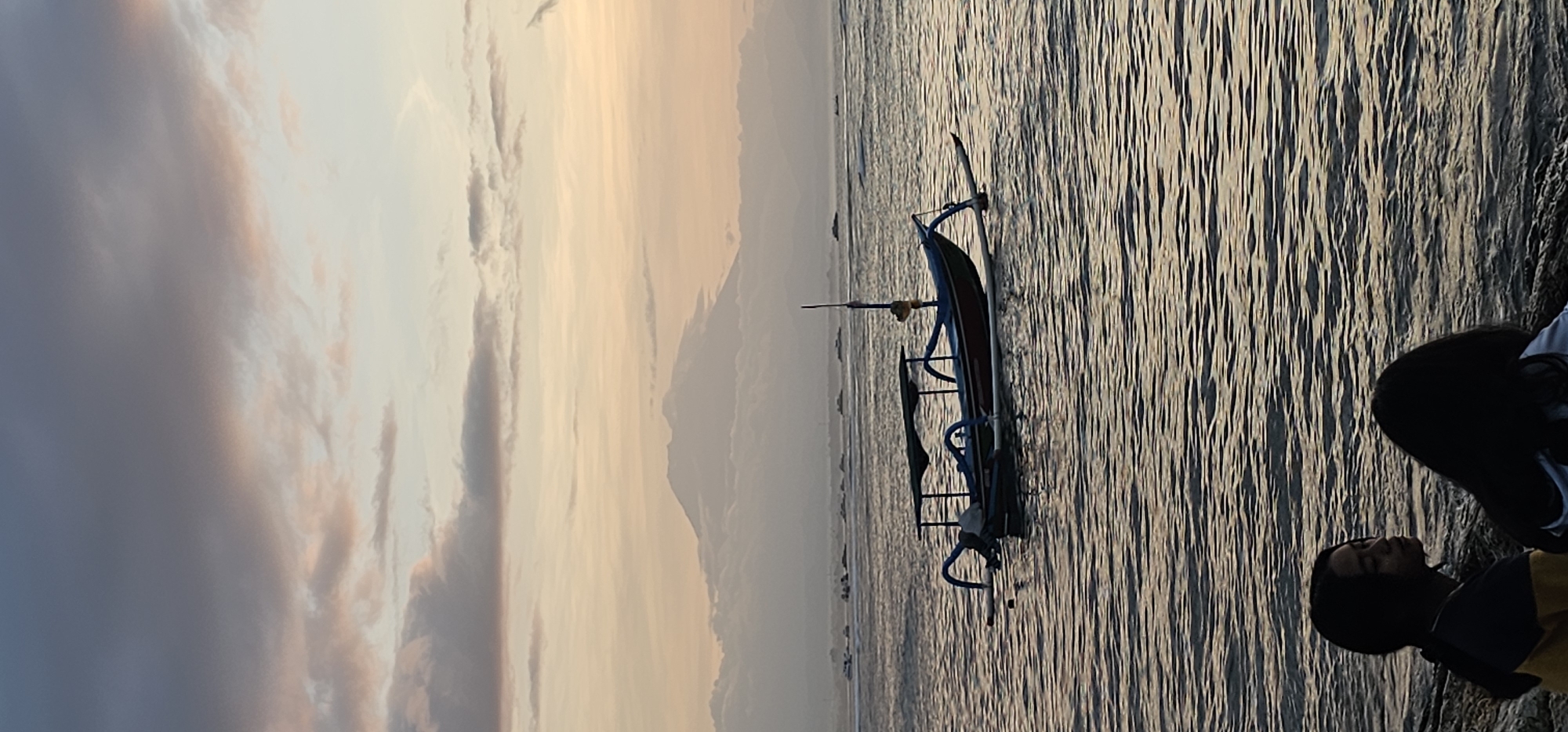 The image depicts a scene on the beach in Denpasar, Indonesia, during either sunrise or sunset. A traditional outrigger boat, with blue and black details, is in the water near the shore. The sky is filled with clouds and shows varying shades of orange and gray. A mountain range is visible in the distance. To the right, a person is sitting, and their face is partially obscured by a mask.