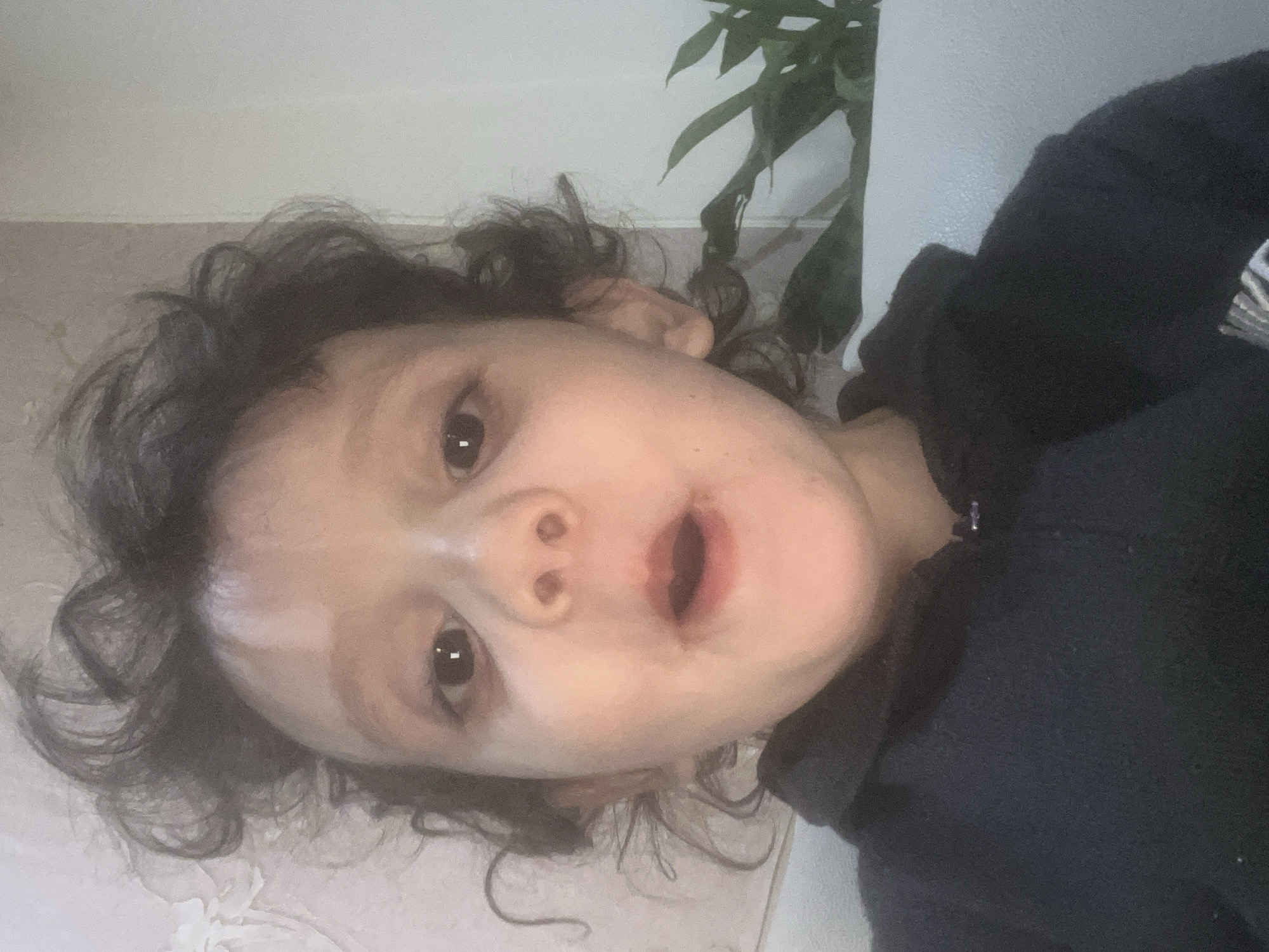 A close-up, tilted shot focuses on a child with curly, dark hair. Their face is the main subject, featuring wide eyes and an open mouth. The child wears a dark-colored hooded sweatshirt. The background is indistinct, showing a wall and part of a green plant. The location and specific environment details are unclear from the image, preventing an exact location identification in Unknown, Unknown.