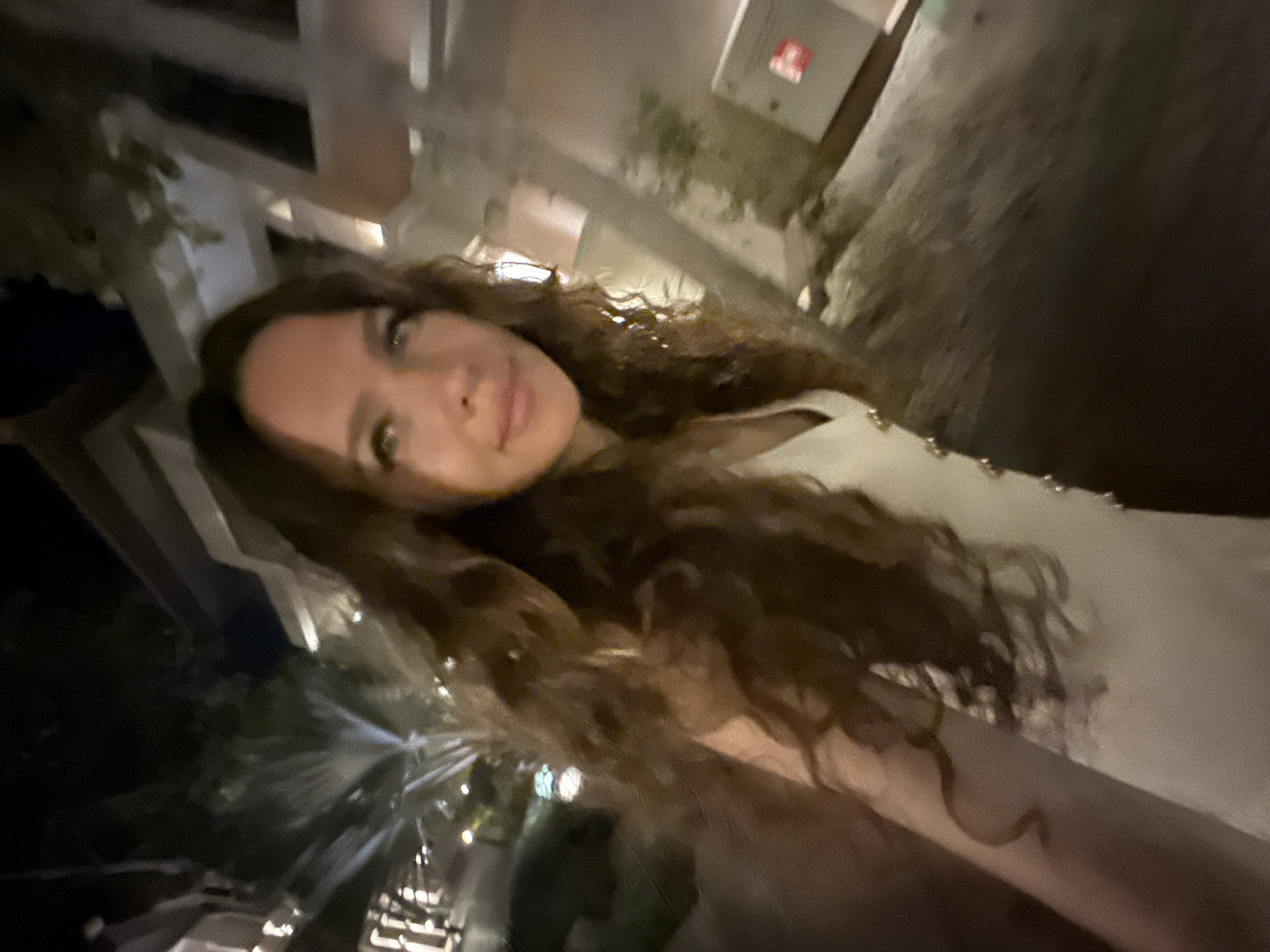 A woman with long, dark, curly hair is taking a selfie in an outdoor, nighttime setting. She is wearing a white dress with embellishments on the bodice. The background appears to be a modern, outdoor area with architectural elements and some foliage. Lights are visible in the background, suggesting an urban environment. The image is slightly blurred, giving it a dynamic feel, possibly indicating movement. The specific location is not identifiable from the image alone.