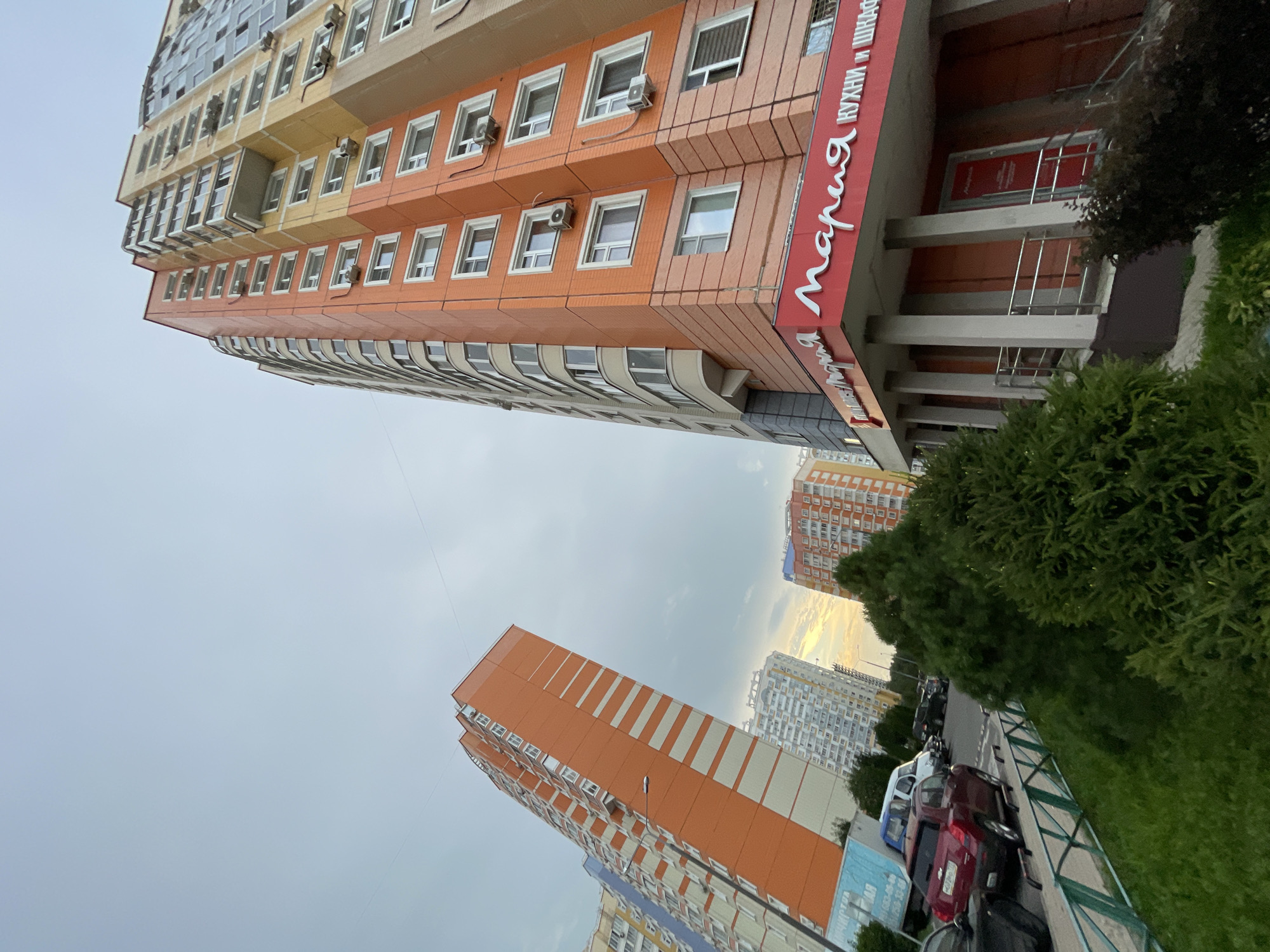 In Moscow, Russia, an upward shot captures several tall apartment buildings under a cloudy sky. The building in the foreground, to the right, is predominantly orange and beige with cream-colored window frames and air conditioning units. A red sign with white Cyrillic lettering that reads "Мария КУХНИ И ШКАФ" (Maria KITCHENS AND CABINETS) is visible at its base, above a red entrance. To the left of this building, another tall apartment building is seen, painted in vibrant orange with white stripes. Further in the background, more apartment buildings with lighter colors are visible. In the lower portion of the frame, parked cars, including a red SUV, a white van, and other vehicles, are seen along a street bordered by a green metal fence and lush green trees and bushes. The overall scene depicts a residential area in a city.
