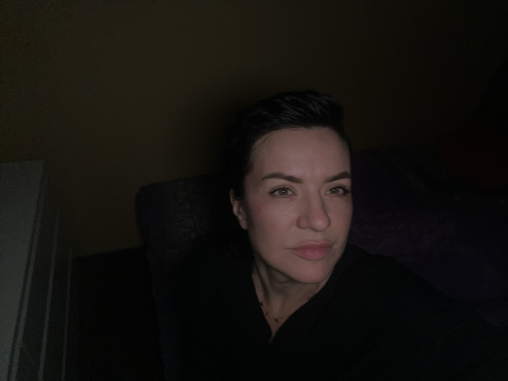 A woman with short dark hair and fair skin is looking directly at the camera in low lighting. She is wearing a dark-colored top and has light-colored lips. The background is dark, suggesting an indoor environment with minimal light sources. There is no clear indication of the location as Belgrade, Serbia, or any specific urban environment.