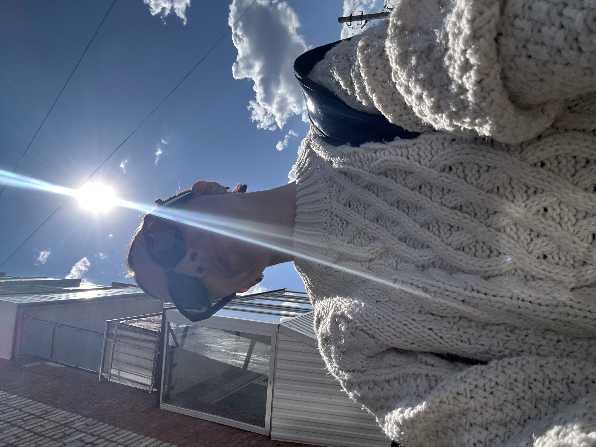 A low-angle photograph captures a person wearing sunglasses and a chunky knit sweater, partially obscuring their face. A black bag strap is visible. The background features a bright blue sky with scattered white clouds and the sun, which produces a lens flare. Part of a building with corrugated metal and glass windows is in the lower-left corner. The setting is inferred to be the city of Votkinsk, Russia.
