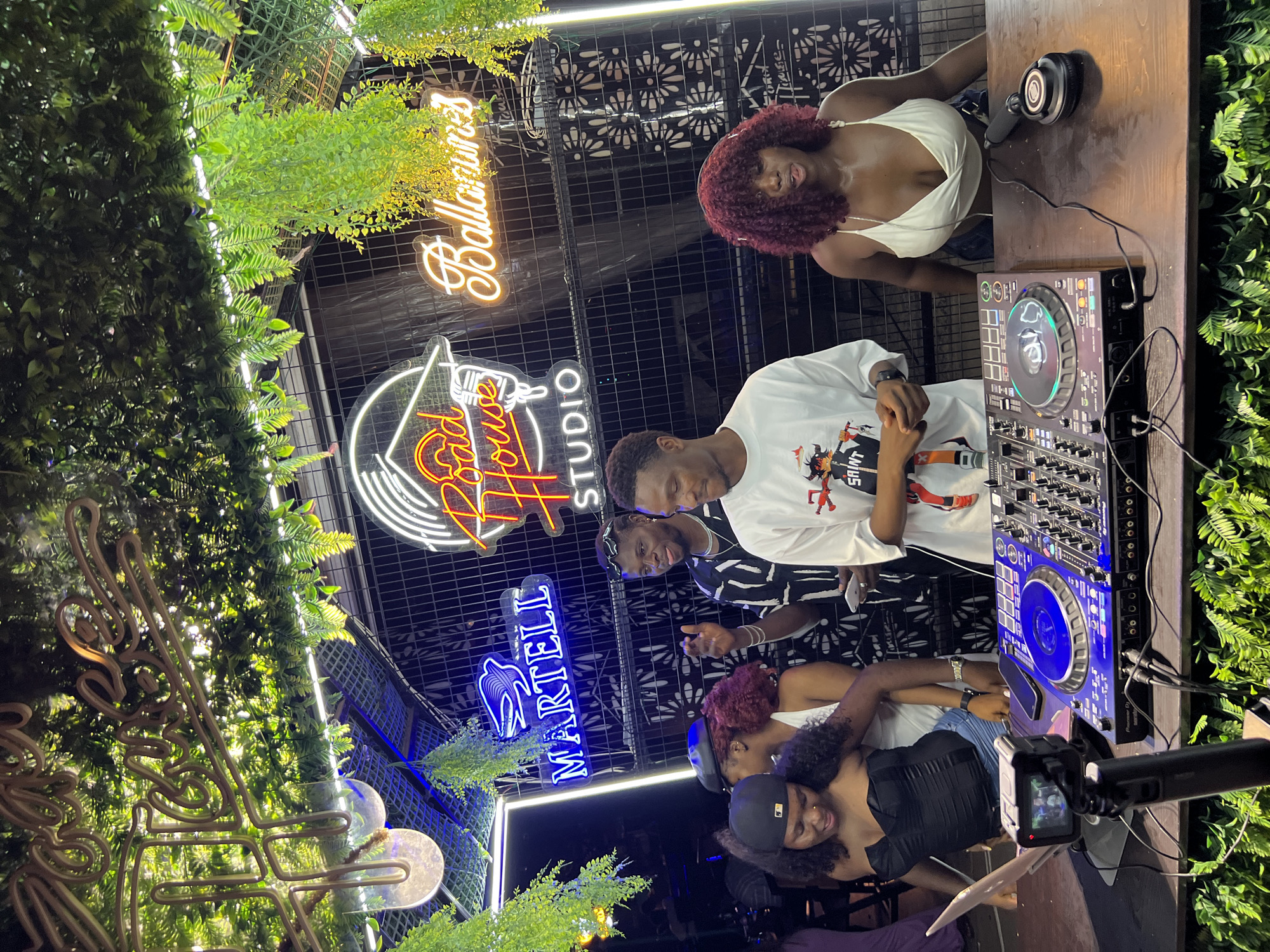 The image depicts an indoor event space, likely a studio or lounge, decorated with abundant hanging greenery and neon signs. One prominent neon sign reads "Road House Studio" in a stylized font, while another, in cursive, says "Ballarunmes." A blue neon sign for "Martell" is also visible. The scene appears to be in Howayi, Nigeria, based on context.
In the foreground, two individuals are positioned behind a DJ console. A woman with dark curly hair and a black corset top is looking down at a laptop and DJ equipment. She is wearing denim shorts. Next to her, a man in a white t-shirt with a graphic print is standing. On the right side of the DJ console, another woman with burgundy curly hair and a white halter top is leaning forward, her mouth slightly open. A pair of headphones rests on the console in front of her.
Behind the DJ, several other people are visible. A man in a patterned shirt is gesturing with his hand, looking towards the DJ. Another man is standing to his right, partially obscured. In the background, a dark fence or screen with a patterned design is visible. The overall atmosphere suggests a lively social gathering, possibly a party or performance event.
