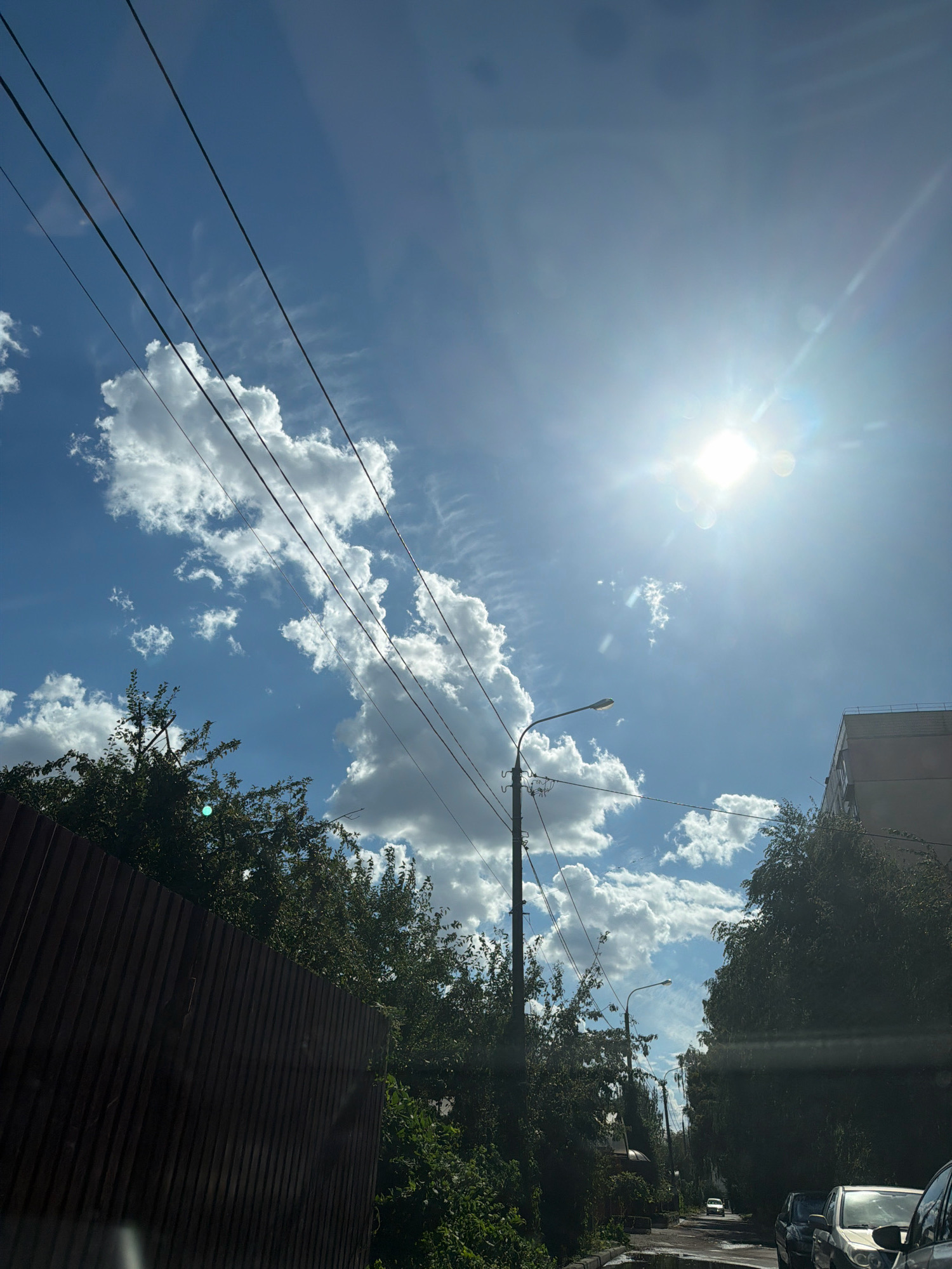 A street scene unfolds under a bright, sunny sky filled with scattered white clouds. The sun is prominently positioned in the upper right quadrant of the image, emitting a strong glare. Power lines crisscross the sky diagonally from left to right. On the left, a tall, dark brown wooden fence lines the foreground. Behind the fence, lush green trees and foliage are visible. A street lamp stands on the left side of the road. In the distance, a multi-story building is partially visible on the right. The road, paved and appearing somewhat wet, curves into the distance, where a white car can be seen. Other vehicles are parked along the right side of the road. The overall impression is an urban environment on a clear day, possibly during the daytime.