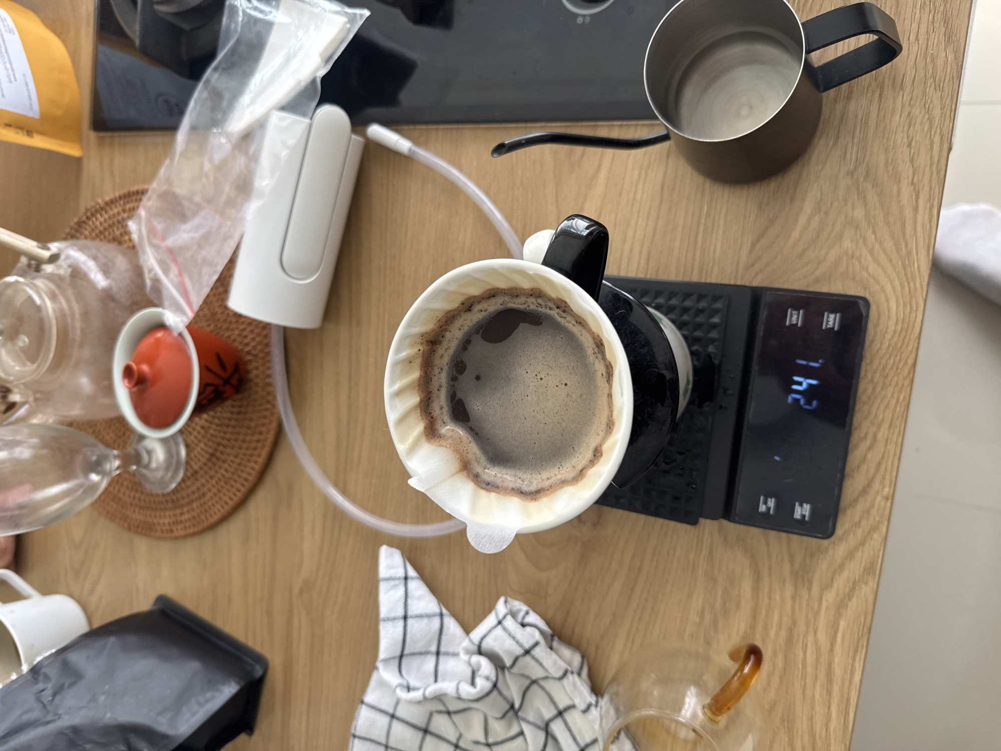 The image shows a close-up, overhead view of a coffee brewing setup on a wooden table. A pour-over coffee maker, with a filter and freshly brewed coffee, is placed on a digital scale displaying "241". Next to it is a gooseneck kettle, a glass carafe, and a small dish with a red lid. In the background, there are various other items including a coffee bag, a teapot, and a small white device. The scene appears to be in a kitchen or dining area, possibly in Kuta Selatan, Indonesia, given the context.