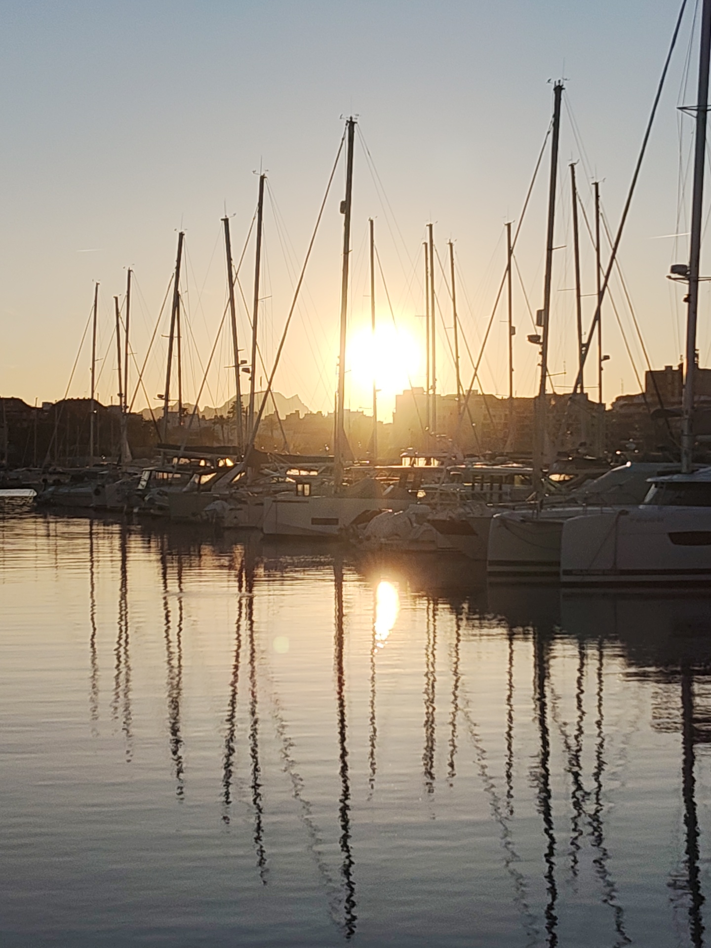 The image shows a harbor scene in Dénia, Spain, at sunset. Several sailboats are moored in the marina. The sun is setting behind the boats and buildings in the background, creating a silhouette effect. The masts of the sailboats are reflected in the calm water of the harbor. There are no people visible in the image.