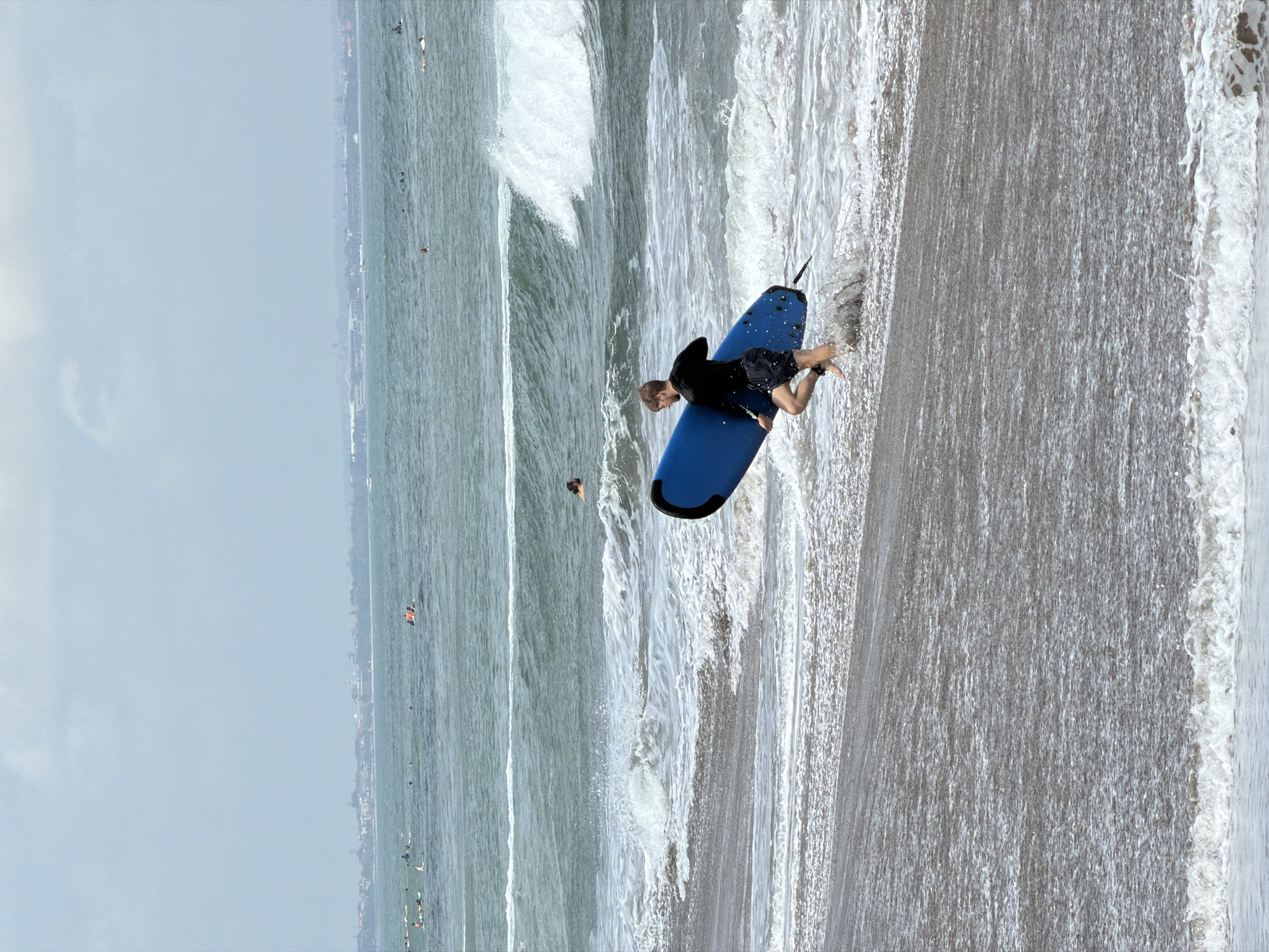 A man is running into the water at the beach with a blue surfboard. He is wearing a black long-sleeved shirt and patterned shorts. The waves are breaking on the shore and there are other surfers in the ocean in the distance. The sky is overcast. The location is the coast of North Kuta, Indonesia.