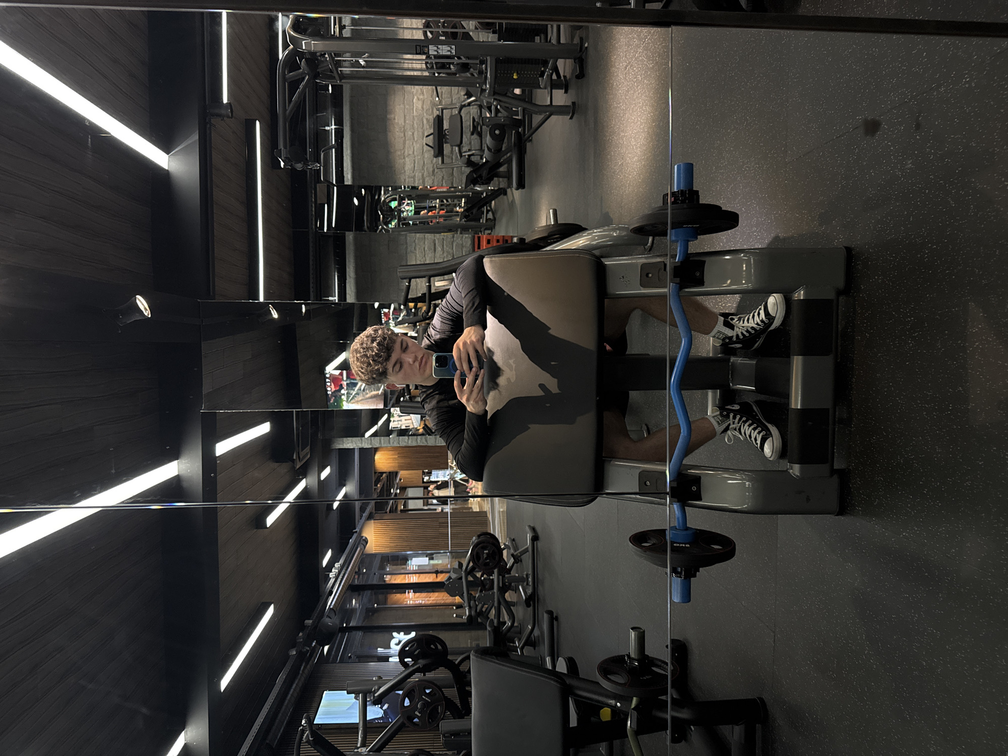A person sits on a leg curl machine in a gym, taking a photo of themselves in the mirror. The gym is well-equipped with various weight machines. The location is likely in Sofia, Bulgaria, although there's no direct evidence in the image to confirm this definitively. The person is wearing a long-sleeved shirt, shorts, and sneakers. The leg curl machine has a blue bar with weights attached.