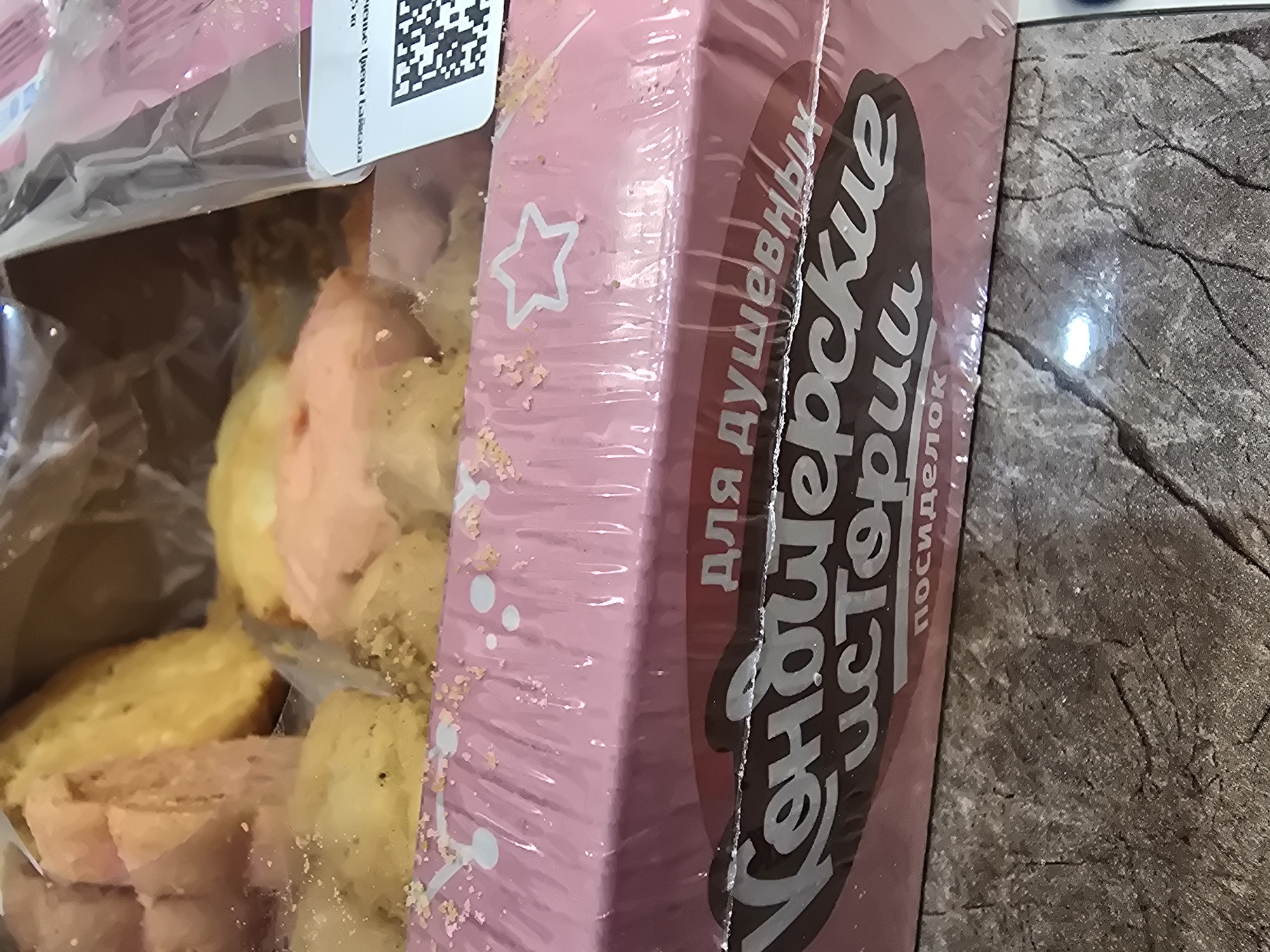 A partially opened box of colorful confectioneries is visible. The box is pink and wrapped in plastic. Inside, several pieces of what appear to be cake or cookies in shades of pink, yellow, and white are arranged. The confectioneries are dusted with fine sugar or sprinkles. A label with Cyrillic text is attached to the right side of the box, along with a QR code. The box sits on a textured surface that resembles a dark stone countertop or table. The lighting suggests an indoor environment. There is no discernible location in the city of Ulan-Ude, Russia.