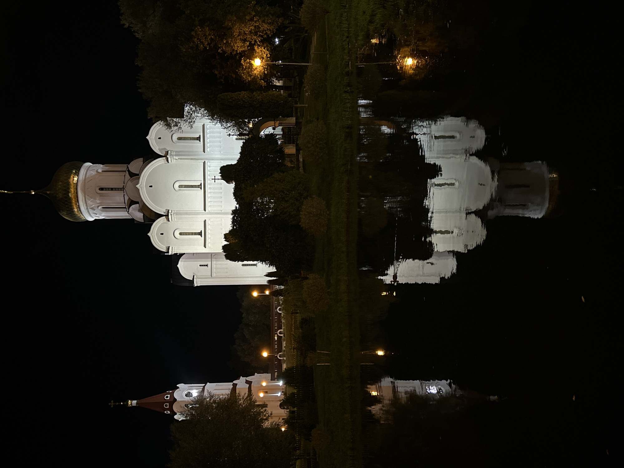 The image depicts a nighttime view of the Holy Spirit Cathedral in Minsk, Belarus, reflected in a body of water. The cathedral, a prominent white building with a golden dome and several smaller white domes, is illuminated by external lights. To the left of the cathedral, a red-roofed bell tower with white accents is also visible. The water in the foreground acts as a mirror, creating a clear reflection of the buildings and the surrounding trees. Several orange streetlights are visible along the pathway in front of the cathedral. The scene is dark, characteristic of nighttime, with the buildings and their reflections being the primary illuminated subjects. There are no people visible in the image.