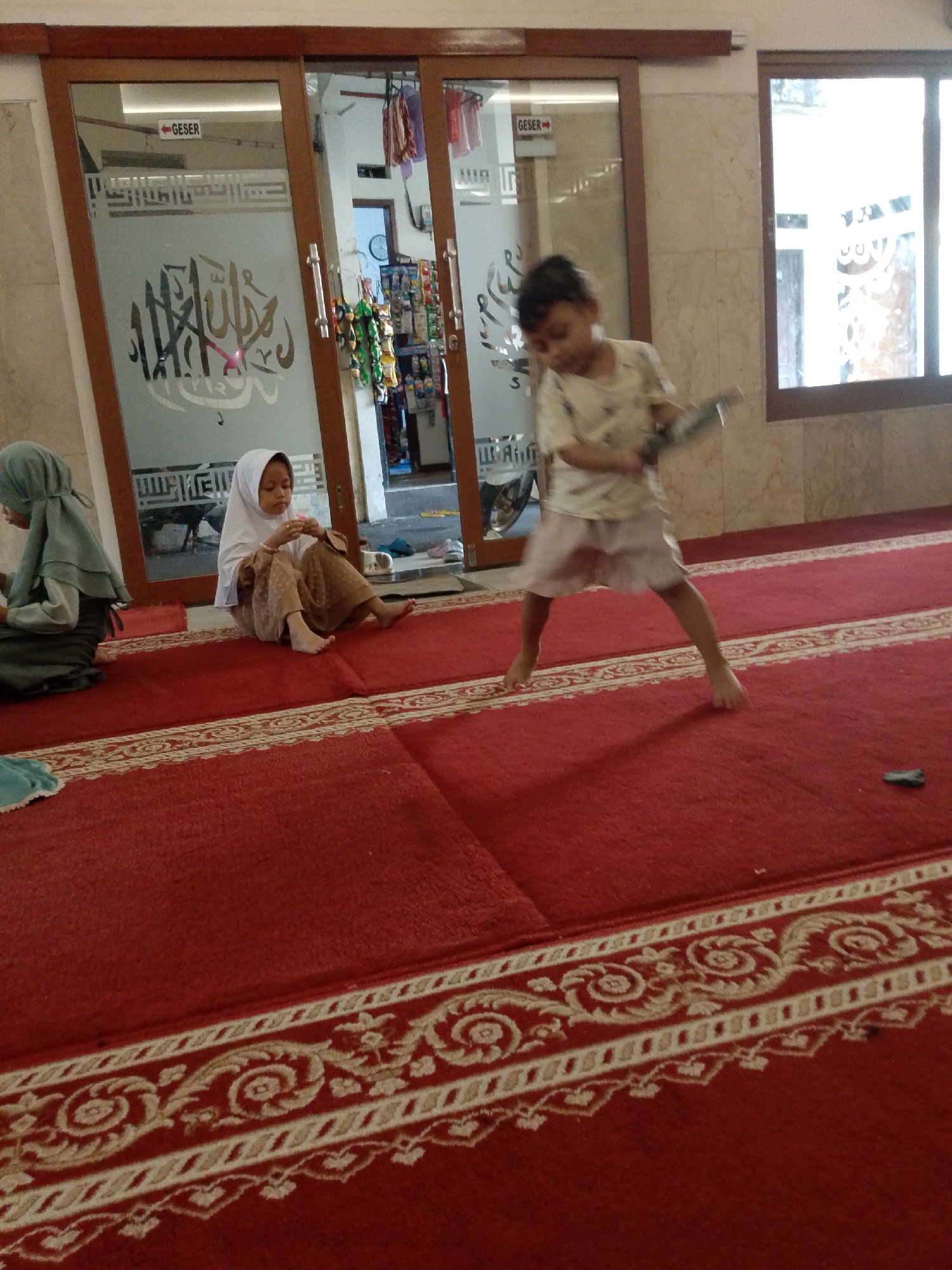The image shows the interior of a mosque in East Jakarta, Indonesia. The floor is covered with a red carpet featuring a decorative border. Two young girls in hijabs are sitting on the carpet. One girl, in a grey hijab, is looking down at something in front of her. The other girl, in a white hijab, is sitting with her legs crossed and is holding a small object. A third child, a younger girl wearing a light-colored patterned shirt and skirt, is standing and appears to be running or playing with a stick-like object. She is blurred due to motion. In the background, there is a glass door with Arabic calligraphy and a sign that reads "GESER" with an arrow, indicating "slide" in Indonesian. Beyond the door, there are shelves with various items, possibly for sale or display. The environment appears to be an indoor prayer area within a mosque.