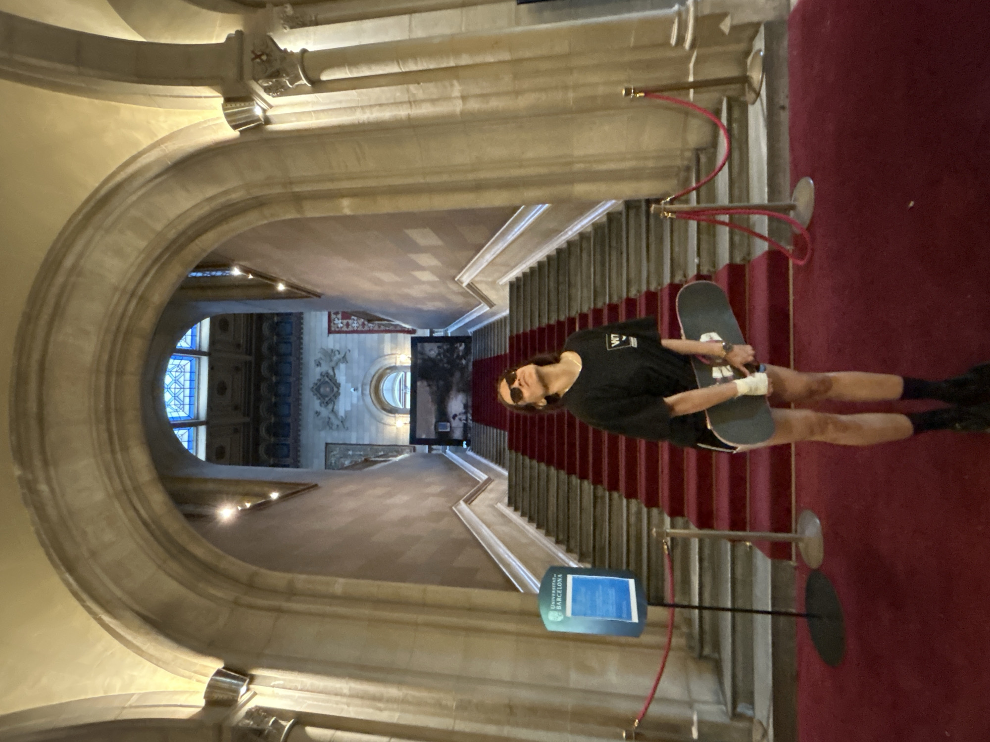 A medium-shot indoor photograph shows a young person holding a skateboard, standing on the right side of a carpeted stairway in an ornate building in Barcelona, Spain. The young person is dressed in a dark shirt and shorts, with dark socks and shoes, and is wearing sunglasses. They hold the skateboard to their right side, at their hip.
The stairway descends in the center of the image, framed by a large archway in the foreground. Red ropes are connected to stands on the right side of the stairway. A sign in the foreground reads "Universitat de Barcelona". Beyond the stairs, an interior view shows arches, windows, and ornate ceilings. The photo is slightly rotated.