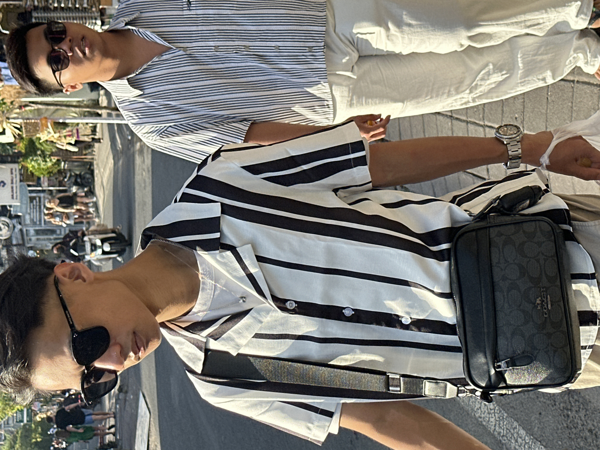 Two young men are walking on a street in North Kuta, Indonesia. The man in the foreground is wearing a black and white striped short-sleeved shirt, a white t-shirt underneath, black sunglasses, and a black crossbody bag. The man behind him is wearing a similar striped shirt and white pants, and also sunglasses. The street appears to be a busy commercial area with shops and other pedestrians in the background, as well as a scooter. The lighting suggests it is daytime.