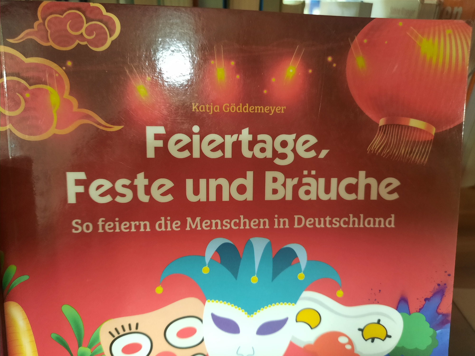 The image shows the cover of a book titled "Feiertage, Feste und Bräuche: So feiern die Menschen in Deutschland" by Katja Göddemeyer. The cover is red and features decorative elements such as clouds and lanterns, as well as cartoonish depictions of masks and facial features. The text is in German. The book cover is likely displayed in a bookstore or library in Rosenheim, Germany, though no specific location within the city can be identified from the image alone.