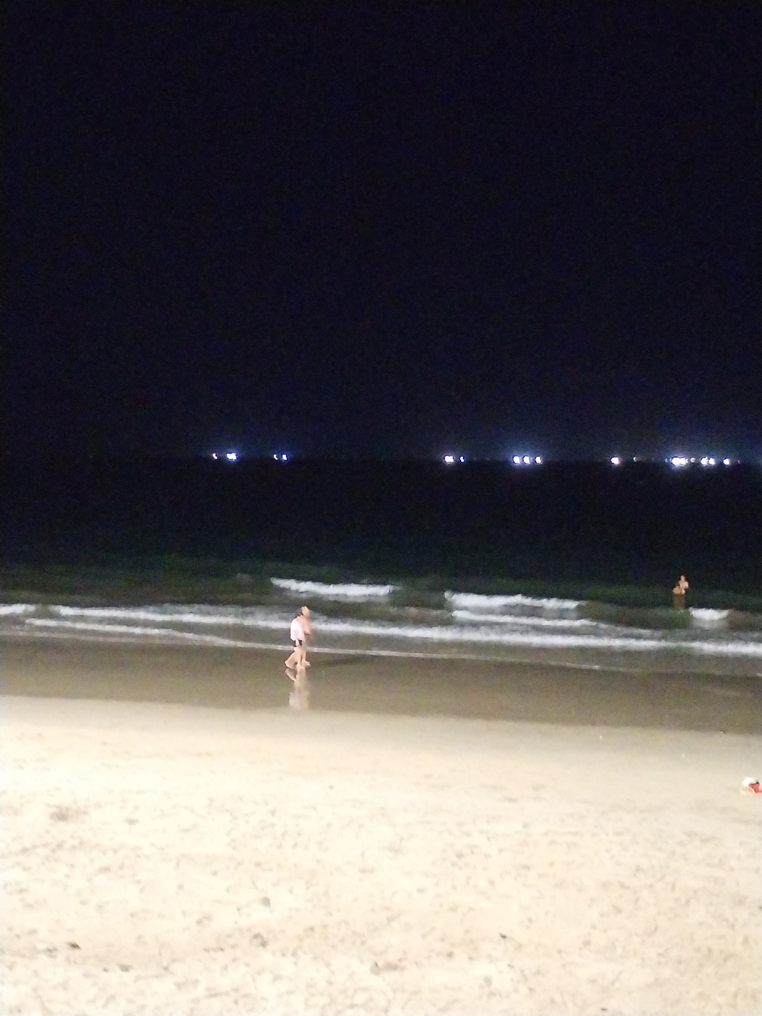 At night, a coastal scene in Nha Trang Ward, Vietnam shows a beach with a dark sky above and the ocean stretching to the horizon. Several bright lights are visible in the distance over the water. Two individuals are present; one is walking along the shoreline, while another appears to be standing in the water. The beach sand is light in color and reflects some light from the water. Waves are visible as they approach the shore.