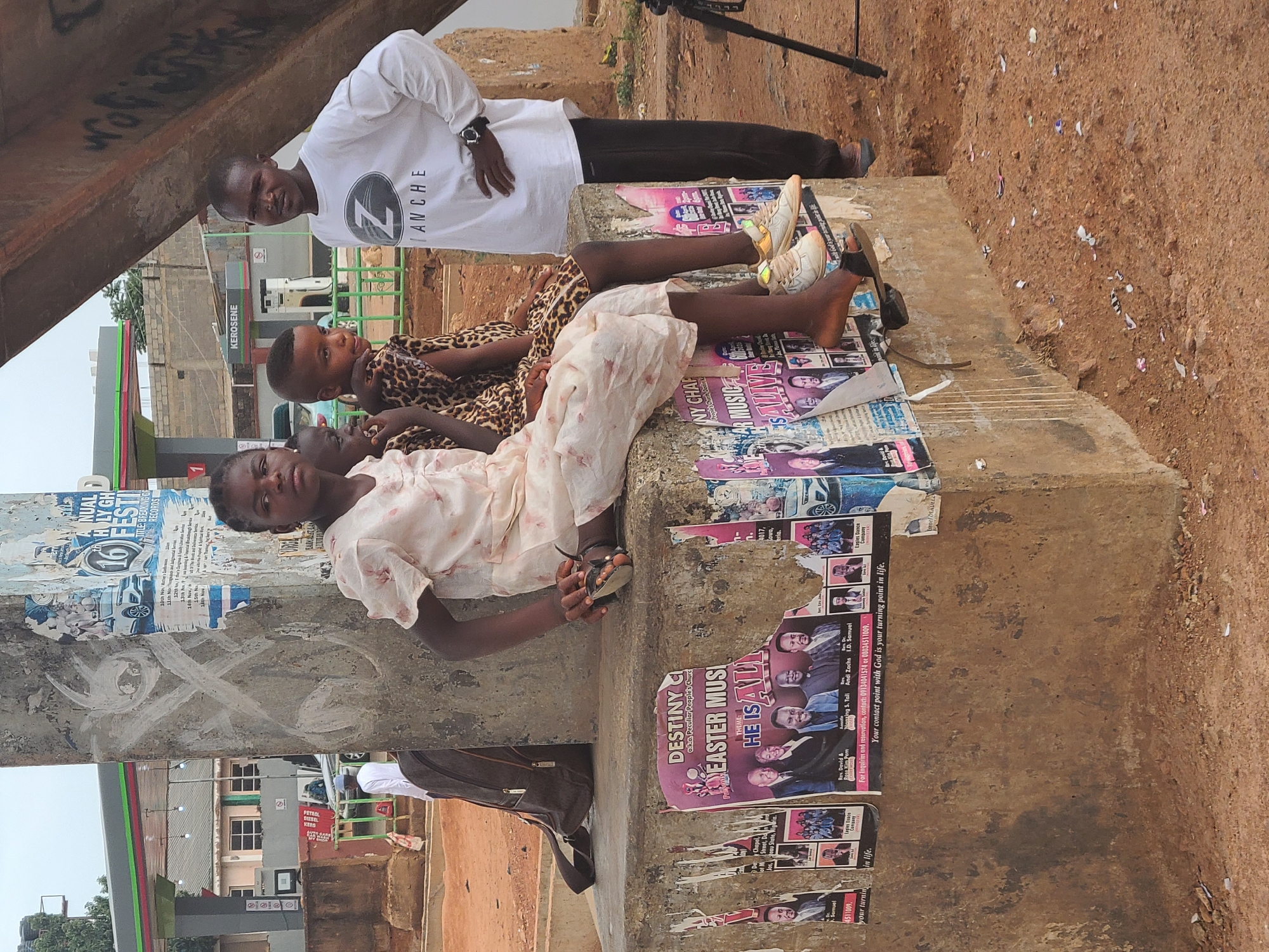 The photograph depicts three individuals in an outdoor setting, potentially Jos, Nigeria, based on the presence of a KEROSENE fuel station sign, posters advertising events from a "Peculiar People's Church," and other contextual cues. An adult male stands wearing a white t-shirt with a "Z ANCHE" logo and black pants. Two younger individuals are lying down on a concrete surface covered in torn posters, with one wearing a leopard print outfit and the other a light-colored dress. The environment appears to be an urban roadside, featuring a building with "Petrol Diesel" signage, a concrete structure possibly part of a building or roadside fixture, and patches of bare ground. The posters on the concrete surface are for various events, including an "Annual Holy Ghost Festival," and feature images of individuals associated with religious organizations. A camera tripod is present in the background.