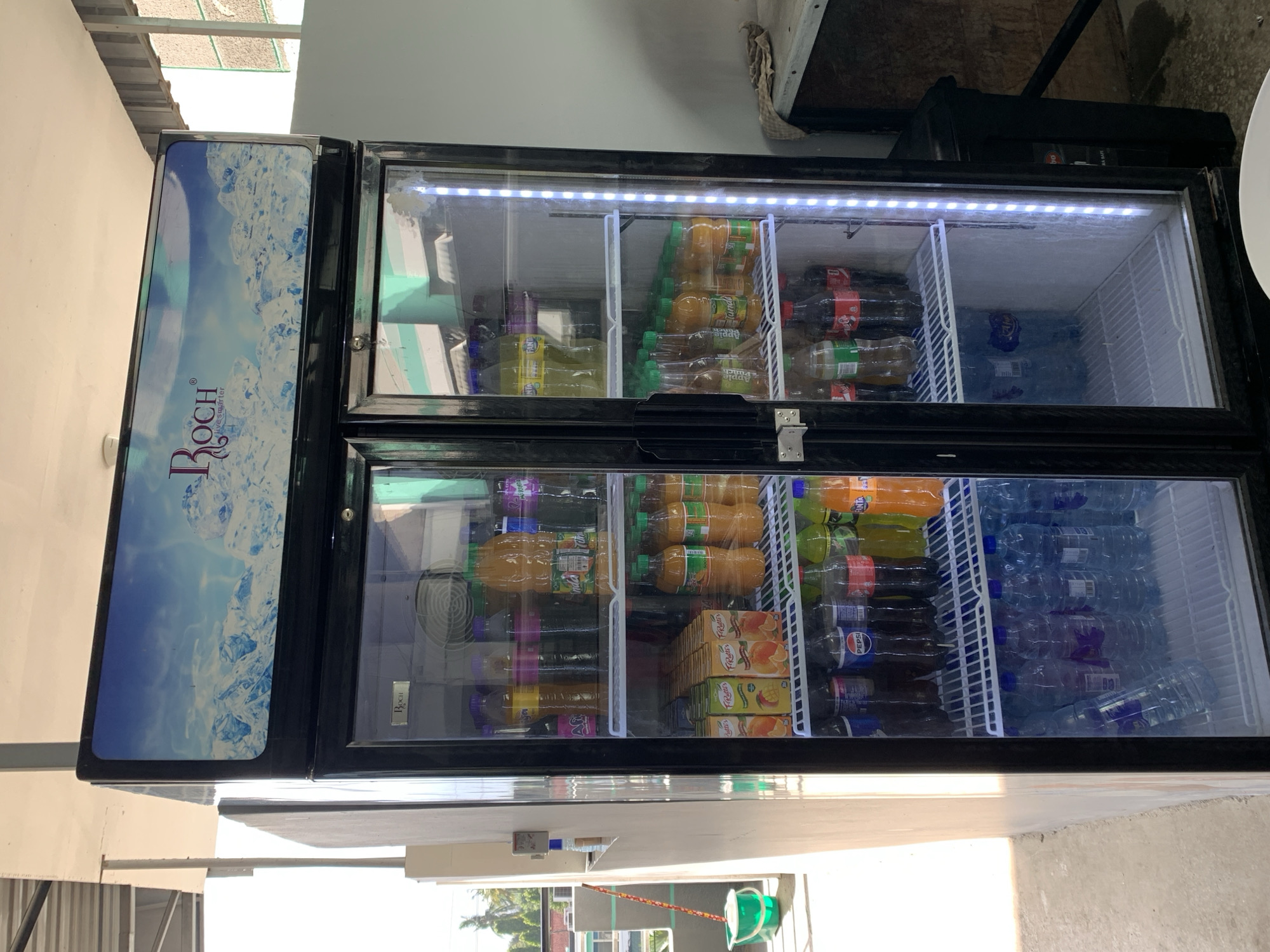 The image shows a well-stocked commercial refrigerator, likely located in Dar es-Salaam, Tanzania, based on the ROCH branding. The refrigerator is filled with various beverages, including bottled water, fruit juices (such as apple punch and mango juice), sodas (Coca-Cola and Pepsi), and other drinks. The bottles are neatly arranged on the shelves within the refrigerator. The refrigerator itself is black and features interior lighting. The surrounding environment includes a building with a corrugated metal roof visible outside.