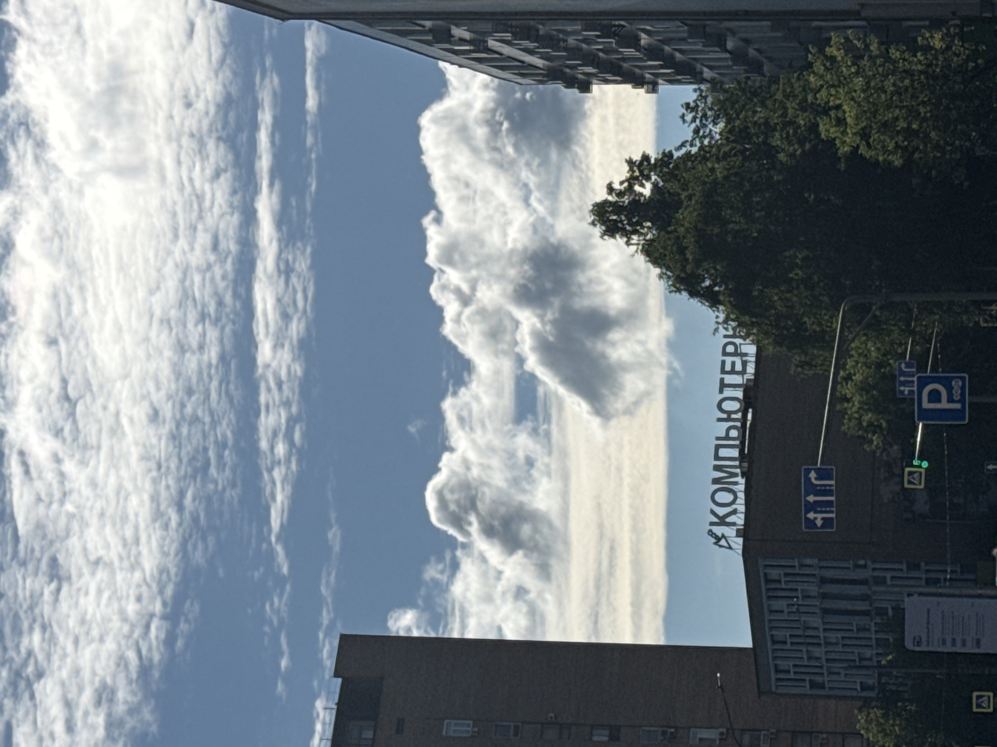 The image depicts a street view in Moscow, Russia, under a partially cloudy sky. On the left, a tall, dark brick building stands prominently. In the center, a building with a patterned facade is visible, topped with a sign that reads "КОМПЬЮТЕРЫ" in blue, illuminated letters. Adjacent to this building, to the right, is another tall building with a grid-like pattern of windows. Lush green trees are scattered throughout the scene, partially obscuring the buildings. Various traffic signs and signals are present, including a blue directional sign with arrows, a green traffic light, and a blue parking sign. The overall impression is an urban environment on a clear day, with a mix of modern architecture and natural elements.