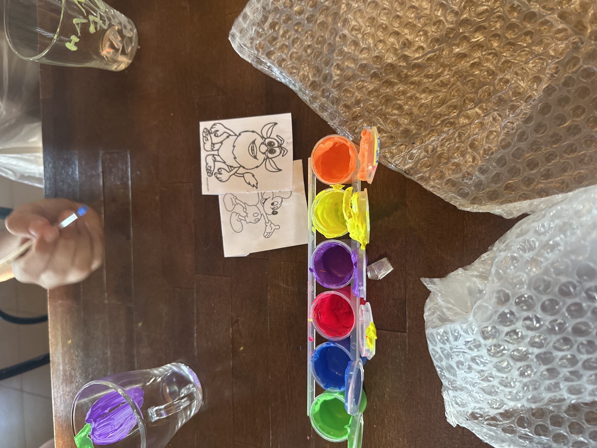 The image shows a wooden table with various objects on it. On the left side, there is a hand holding a stick, presumably a paintbrush. There are two glasses, one with green and yellow writing on it and another with a purple flower painted inside. There are also two black and white drawings of cartoon-like creatures. A container of colorful paints is arranged in a vertical stack. To the right of the objects are large pieces of bubble wrap.