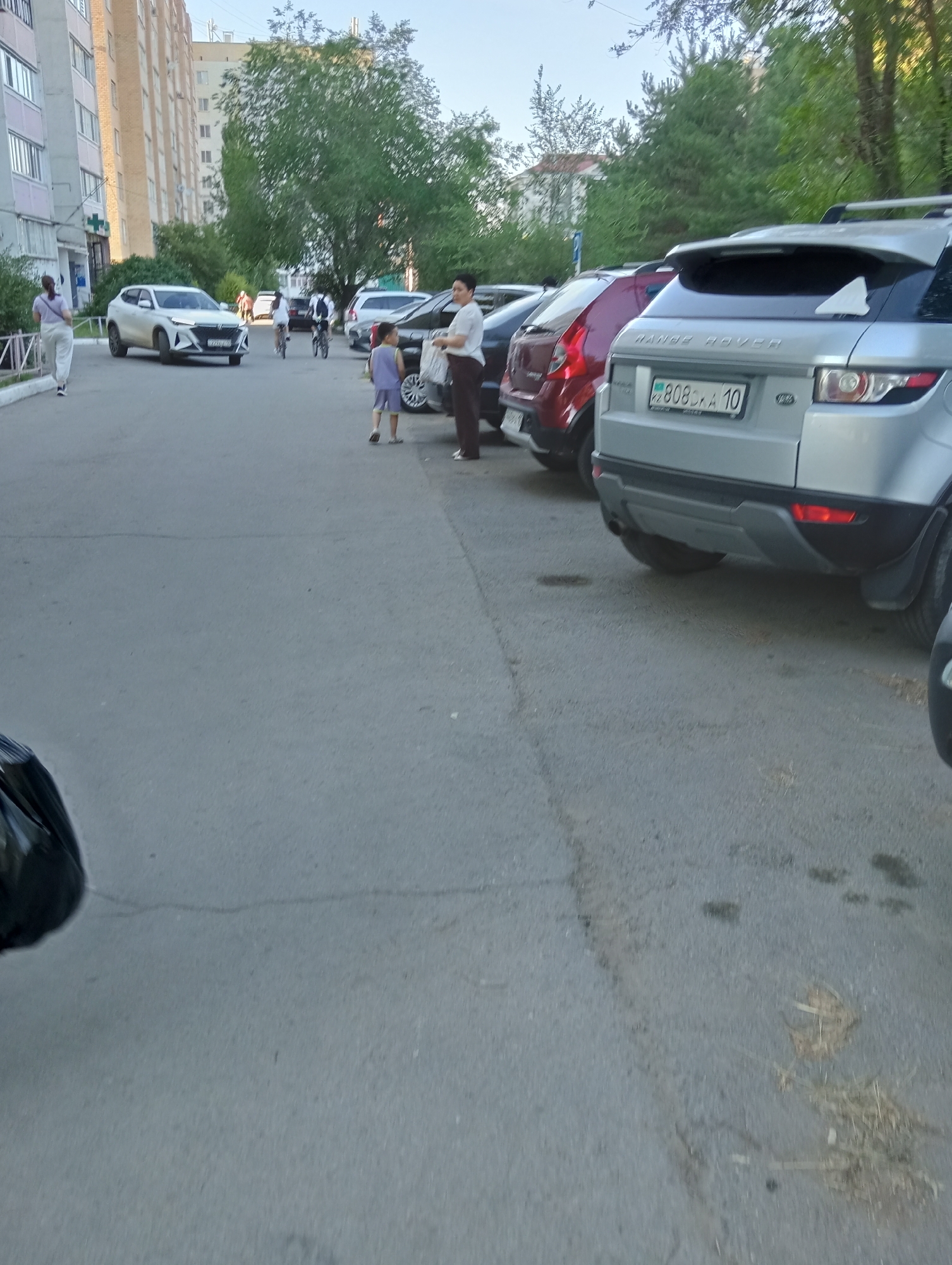 The image captures a street scene in what appears to be a residential area. The foreground is dominated by a grey asphalt road, with some dried grass and debris scattered on its surface. In the background, several cars are parked. One prominent vehicle is a silver Range Rover with a license plate reading "KZ 808 KXA 10". Adjacent to it are other vehicles, including a red car and a white SUV.
Several people are visible in the image. On the left side, a woman in a light-colored top and pants walks away from the camera. Further into the scene, a woman in a white top and dark pants stands near a child dressed in a light purple outfit. In the mid-ground, a few individuals are seen on bicycles, suggesting recreational activity or commuting.
The setting includes apartment buildings lining the street, indicating an urban or suburban environment. Tall green trees with dense foliage are also present, casting shadows and contributing to the overall ambiance. The lighting suggests it is daytime, likely late afternoon or early evening, given the warm light and shadows. The mood seems ordinary and relaxed, with people going about their day. No specific event is clearly indicated, but the presence of pedestrians and cyclists suggests a typical day.