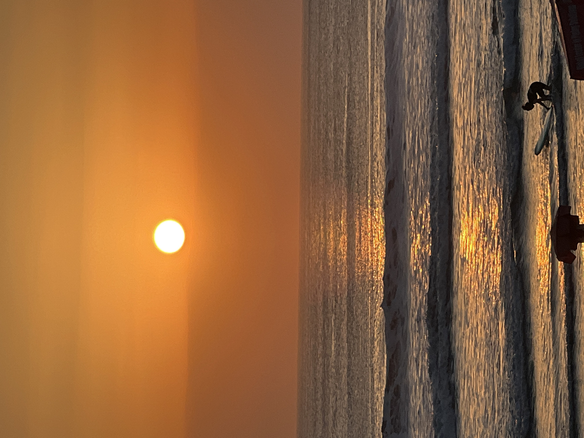 The image shows a sunset over the ocean in Kuta, Indonesia. The sun, a bright orange disc, is positioned in the upper center of the frame, casting a golden reflection on the water. The sky is a gradient of warm orange and yellow hues. Several waves are breaking on the shore, with white foam visible. In the foreground, a person is kneeling on a surfboard in the shallow water, facing away from the camera. The person appears to be a surfer getting ready to catch a wave. There is a dark, indistinct object in the lower right corner which may be a portion of another surfboard or beach equipment. The scene conveys a sense of tranquility and the beauty of a tropical sunset.