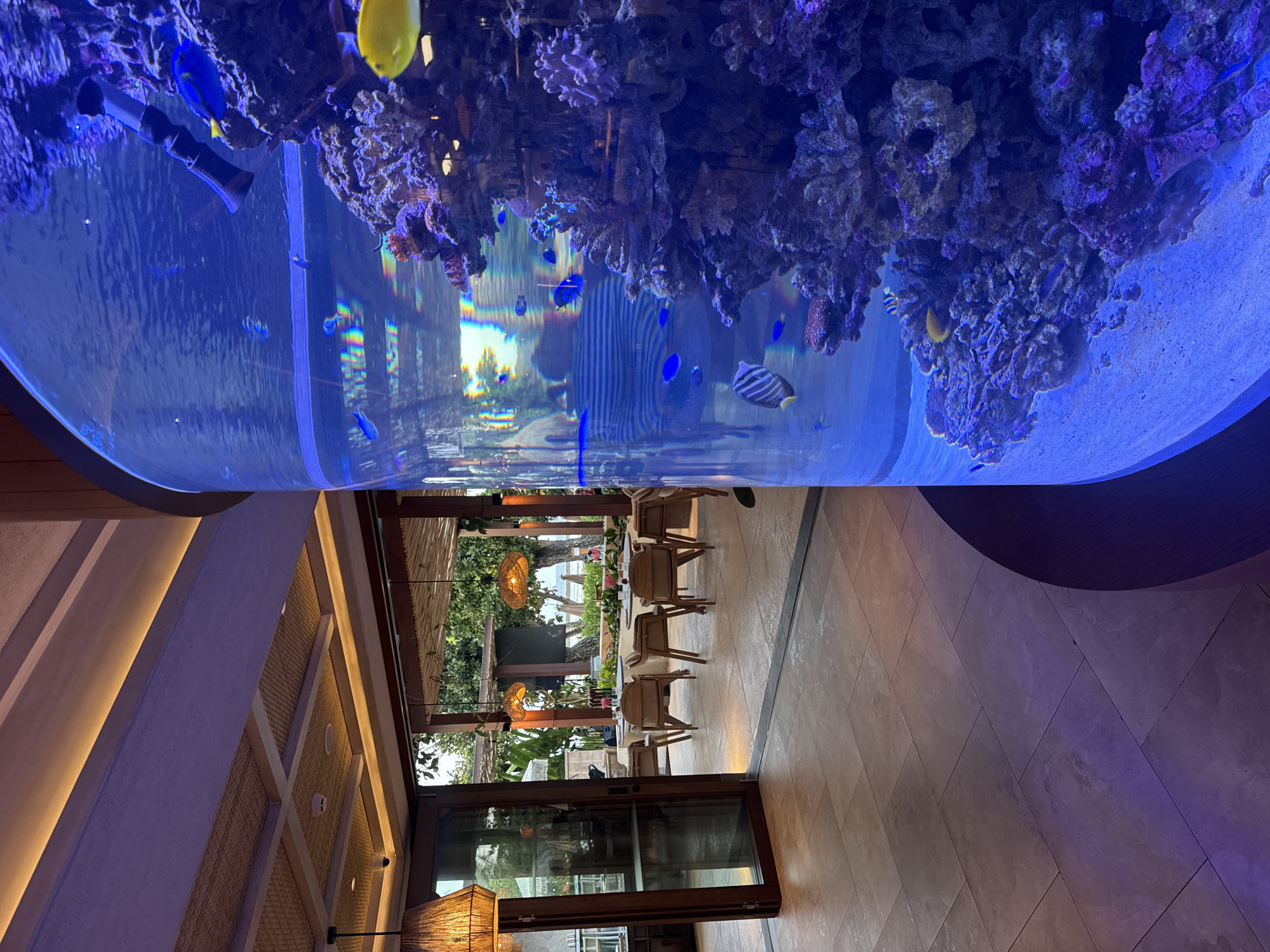 This is a photograph of the interior of a restaurant in Kuta Selatan, Indonesia, with a large cylindrical aquarium dominating the right side of the frame. The aquarium is filled with clear blue water, marine life, and coral structures. Several fish are visible, including a prominent yellow tang and a blue tang. The restaurant interior features tiled flooring and a ceiling with woven accents and integrated lighting. Through an open doorway to the left, a dining area is visible with round tables set for meals. Beyond the dining area, an outdoor setting with trees and a beach can be seen in the distance. The overall ambiance suggests a tropical, upscale dining establishment.