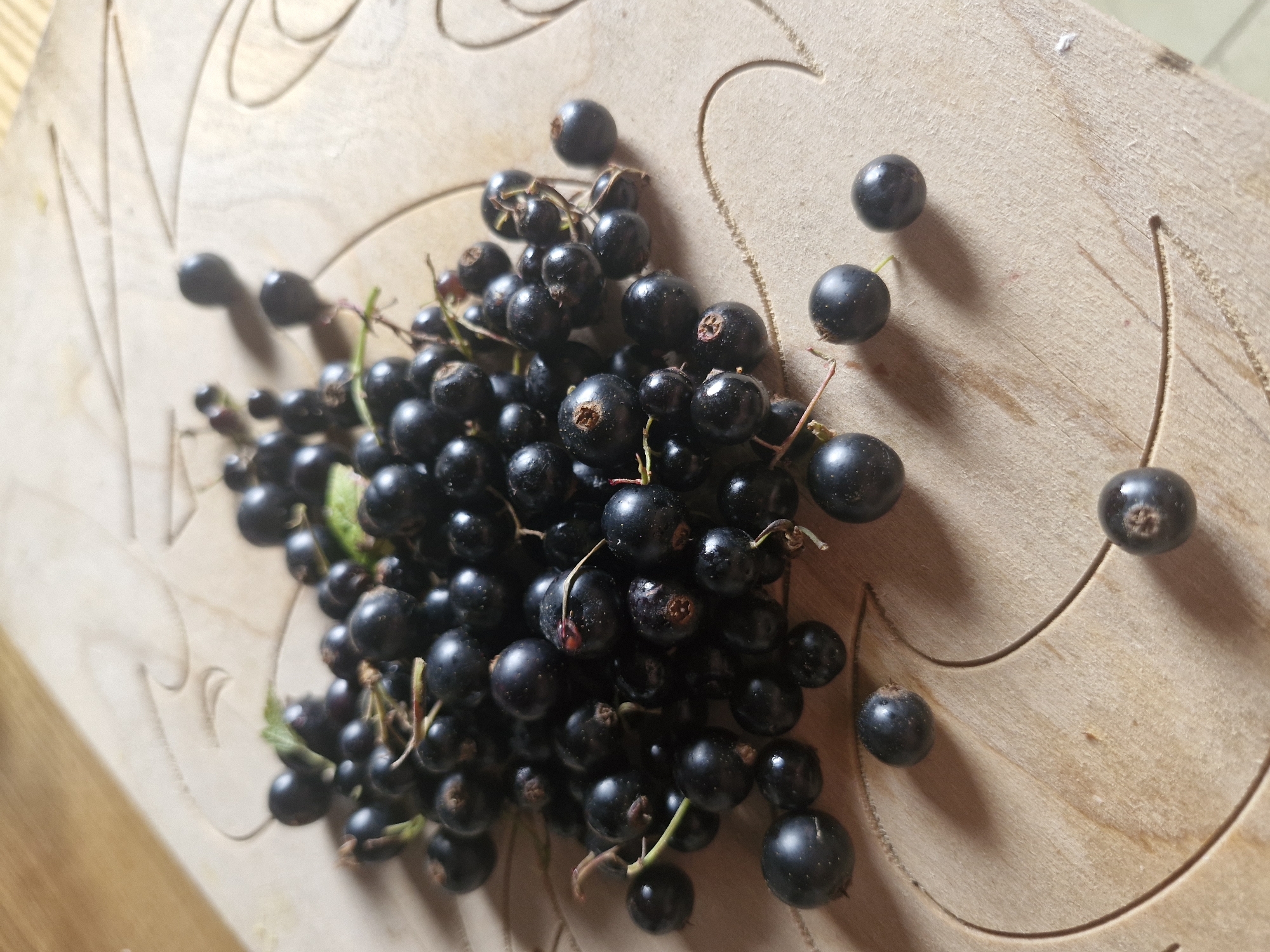 A pile of fresh, dark black currants is scattered across a light-colored wooden surface. The currants vary in size, with some clustered together and others spread out individually. The wooden surface appears to be a cutting board or tabletop with decorative carvings. A few small green leaves are interspersed among the currants. The lighting suggests an indoor setting, possibly a kitchen. The image does not provide any information that could place it in the city of Ķekava, Latvia, or any other specific location.