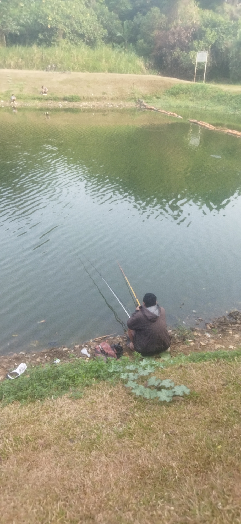 The image depicts a serene outdoor scene, likely in Depok, Indonesia, featuring a calm body of water. In the foreground, a person is seated on the grassy bank, engaged in fishing. Two fishing rods are extended into the water, suggesting an attempt to catch fish. The individual is dressed in casual attire, with a hooded jacket. The surrounding environment includes lush green vegetation along the water's edge and a gentle slope of dry grass leading away from the bank. In the background, trees and shrubs form a natural backdrop, and a signpost is visible near the water's edge. A small group of people and some bicycles are visible in the distance on the far bank. The overall atmosphere is peaceful and relaxed.