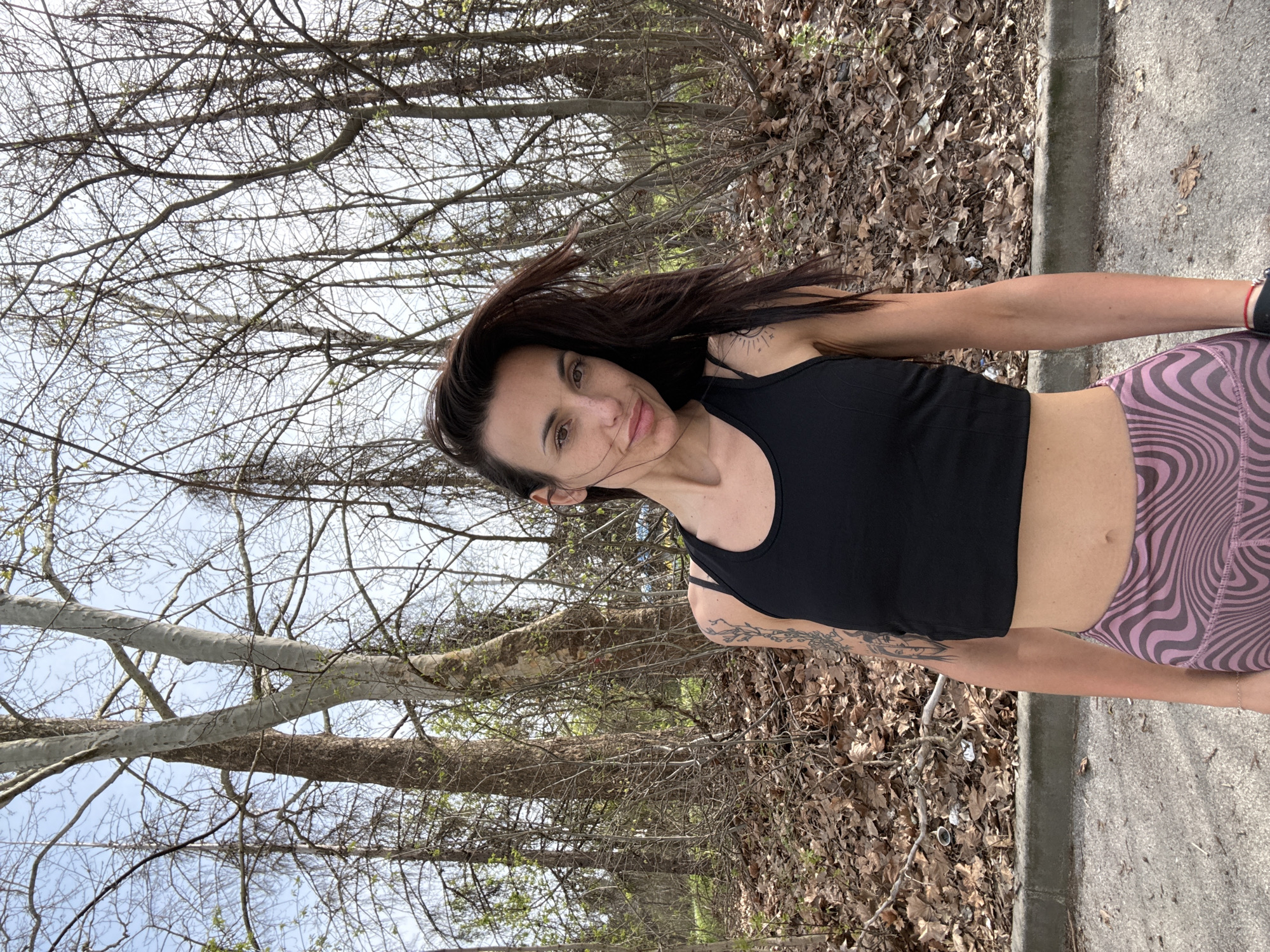 A woman stands outdoors on a paved surface adjacent to a wooded area. She wears a black tank top and patterned leggings. The background features trees with bare branches and fallen leaves. The location's connection to Pleven, Bulgaria, is not ascertainable from the visual information.