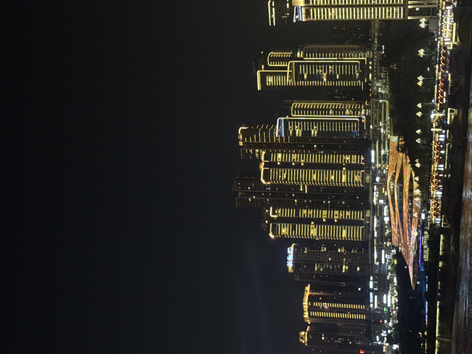 A nighttime cityscape view captures a cluster of modern skyscrapers with illuminated windows and exterior lighting, creating a vibrant glow against the dark sky. The buildings are densely packed, showcasing a range of heights and architectural styles. In the foreground, a body of water reflects the city lights, and several boats or barges are visible, adorned with their own illumination. A multi-tiered illuminated structure with a wave-like design is prominent in the mid-ground, adding to the overall spectacle of the urban night scene. The image is set in the Yuzhong District, China.