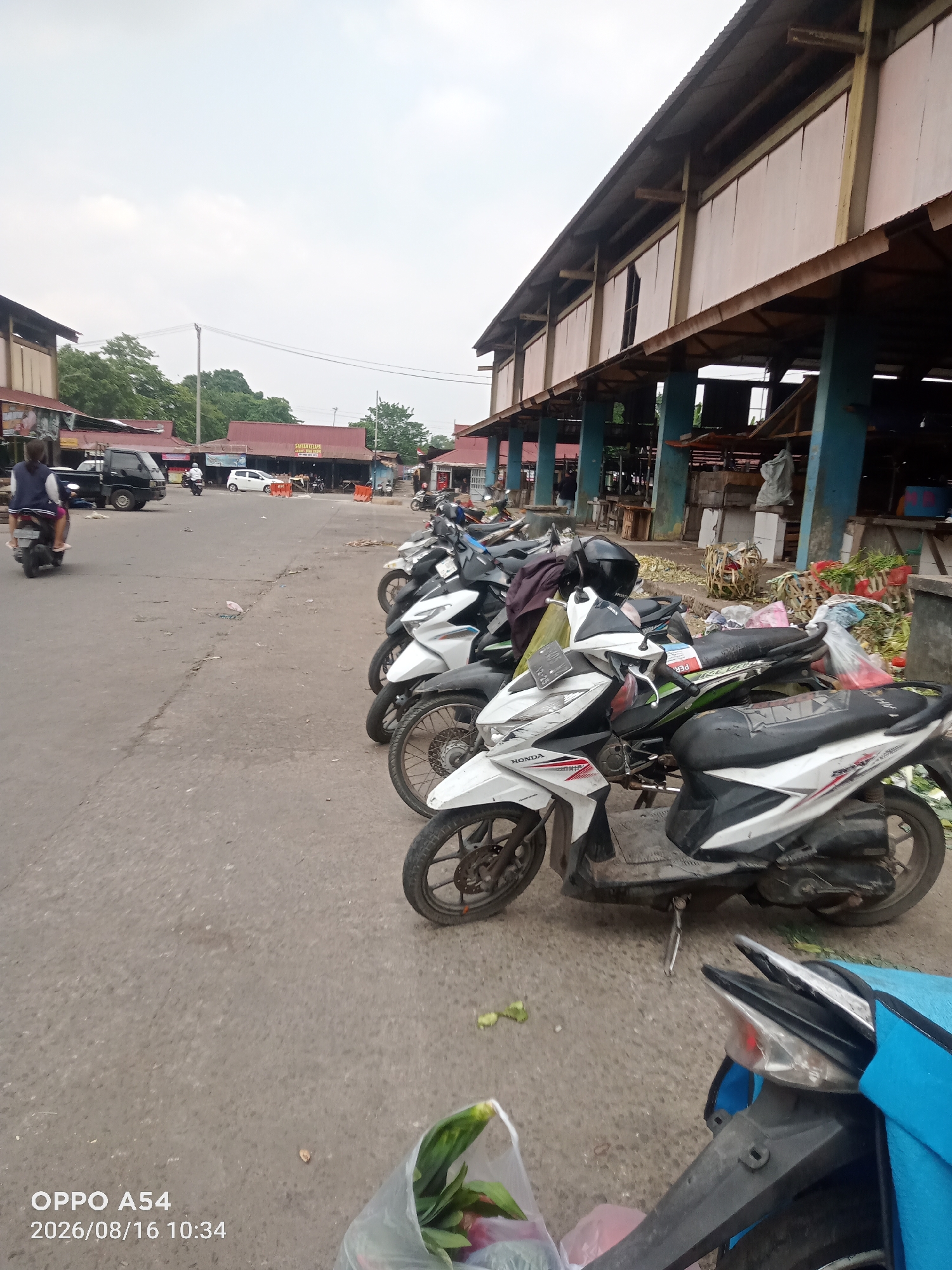 A street scene in the city of Jambi, Indonesia, shows a row of parked motorcycles and scooters on the right side of the frame, some with their headlights and taillights illuminated. In the background, a long, covered market structure with a corrugated metal roof is visible, supported by blue pillars. Several stalls within the market appear to be open, with goods displayed. On the left side of the street, a black pickup truck is parked, and a person is riding a scooter away from the camera. Further down the street, other vehicles, including a white car, are present. The ground is paved with asphalt, and there are scattered items on the surface, possibly debris or produce. The sky is overcast, suggesting a cloudy day. A digital timestamp "2026/08/16 10:34" and "OPPO A54" are visible in the bottom left corner, likely from the camera that captured the image.