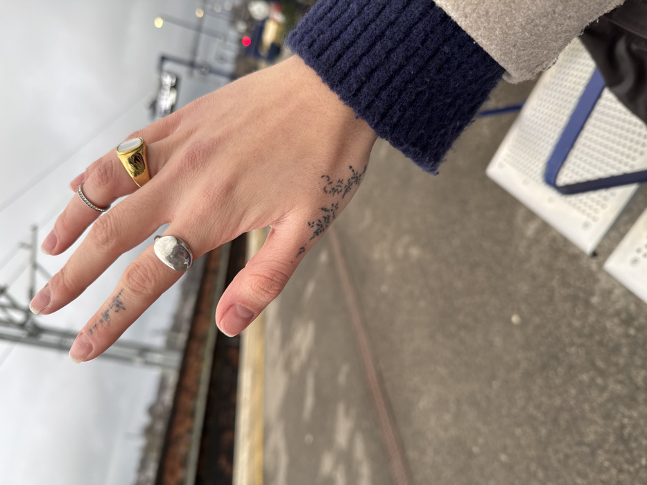 A pale hand adorned with three rings is in focus. A gold ring with a possible mother-of-pearl inset is on the ring finger, a thin silver band on the middle finger, and a silver signet ring on the index finger. Tattoos are visible on the thumb and along the side of the hand, near the wrist. A blue knit sleeve peeks out from a grey or beige coat. The hand is positioned over a grey platform with the edge of a train track visible in the background.