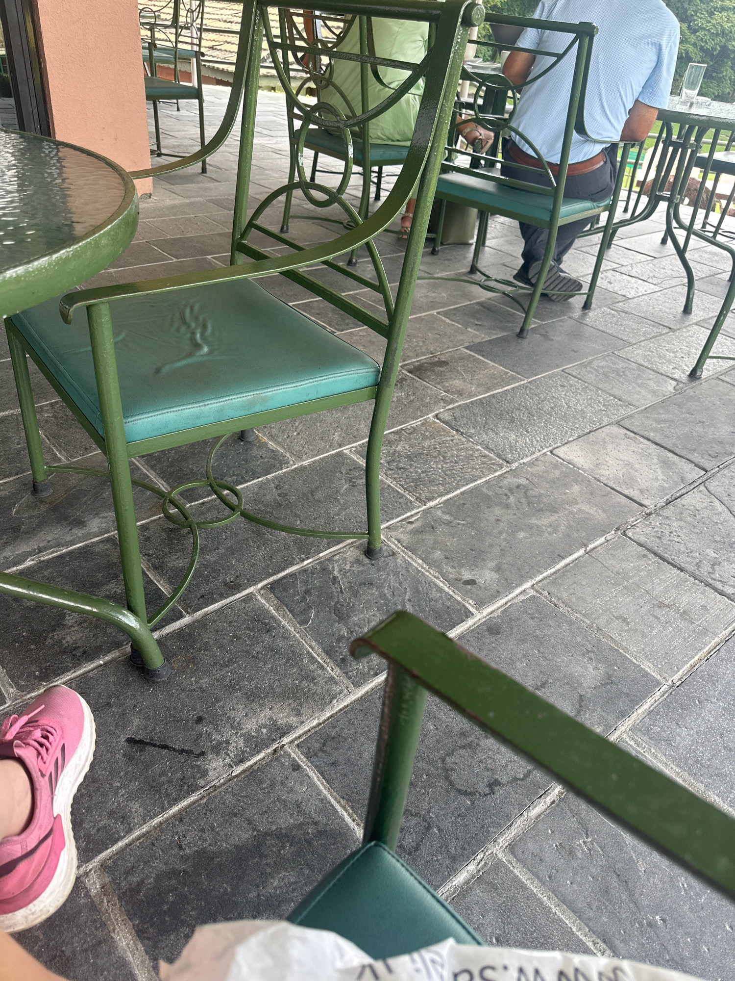 An overhead outdoor patio scene depicts several green metal chairs and tables. The flooring is a dark gray stone tile laid in a grid pattern. In the foreground, a person's right foot wearing a pink athletic shoe is visible. Further back, a person in a light blue collared shirt and dark pants is seated at a table, their back to the camera. Another person, only partially visible, is seated at a table to the left, wearing a light green garment. The overall atmosphere suggests a casual outdoor dining or seating area. The location is Kageshwori Manohara, Nepal.