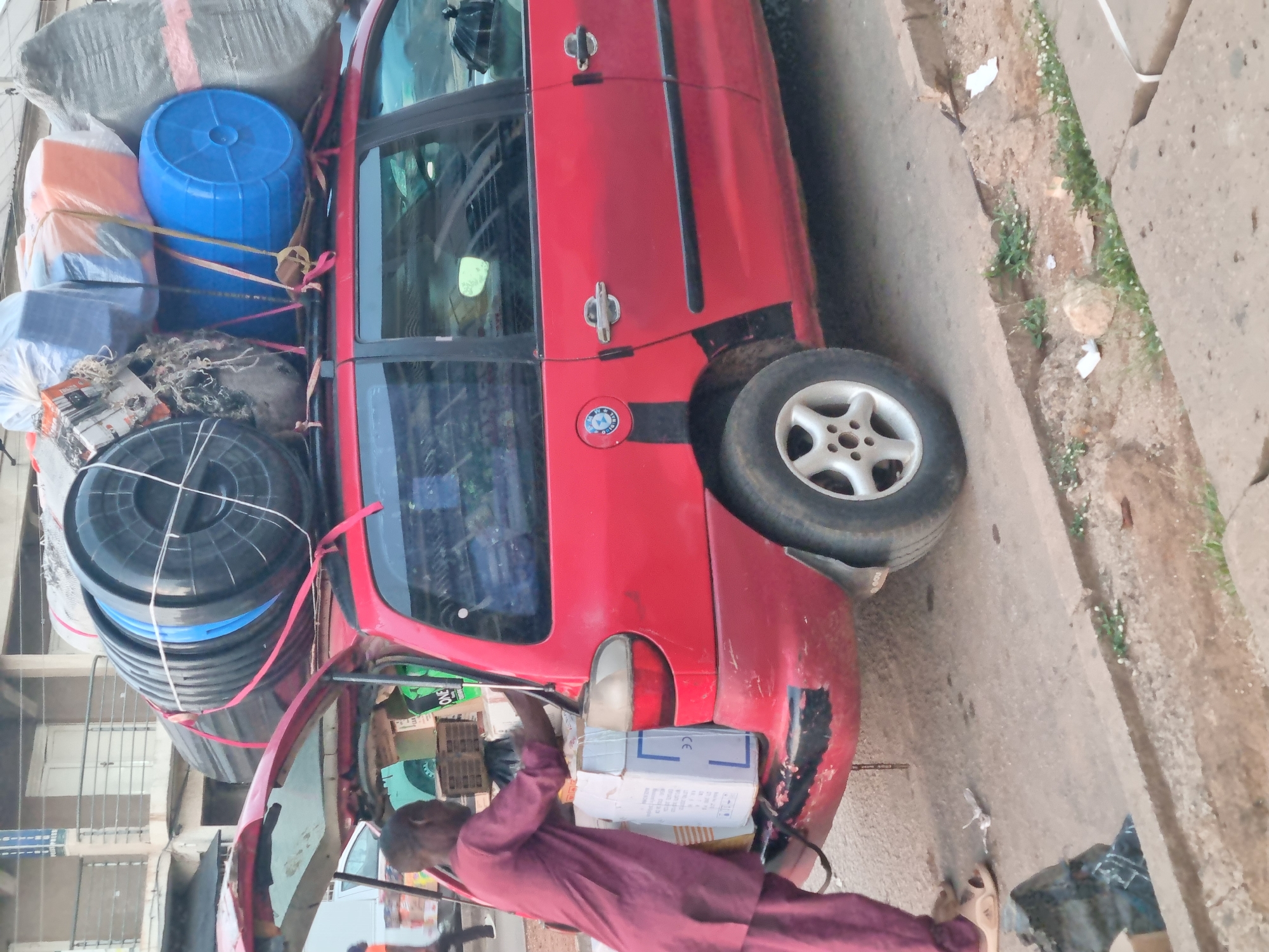 In Jos, Nigeria, a red minivan is packed with an excessive amount of cargo, including barrels and wrapped bundles, strapped to its roof. A person in a burgundy robe is loading boxes into the vehicle's trunk. The car is parked on a street next to a sidewalk and gutter.