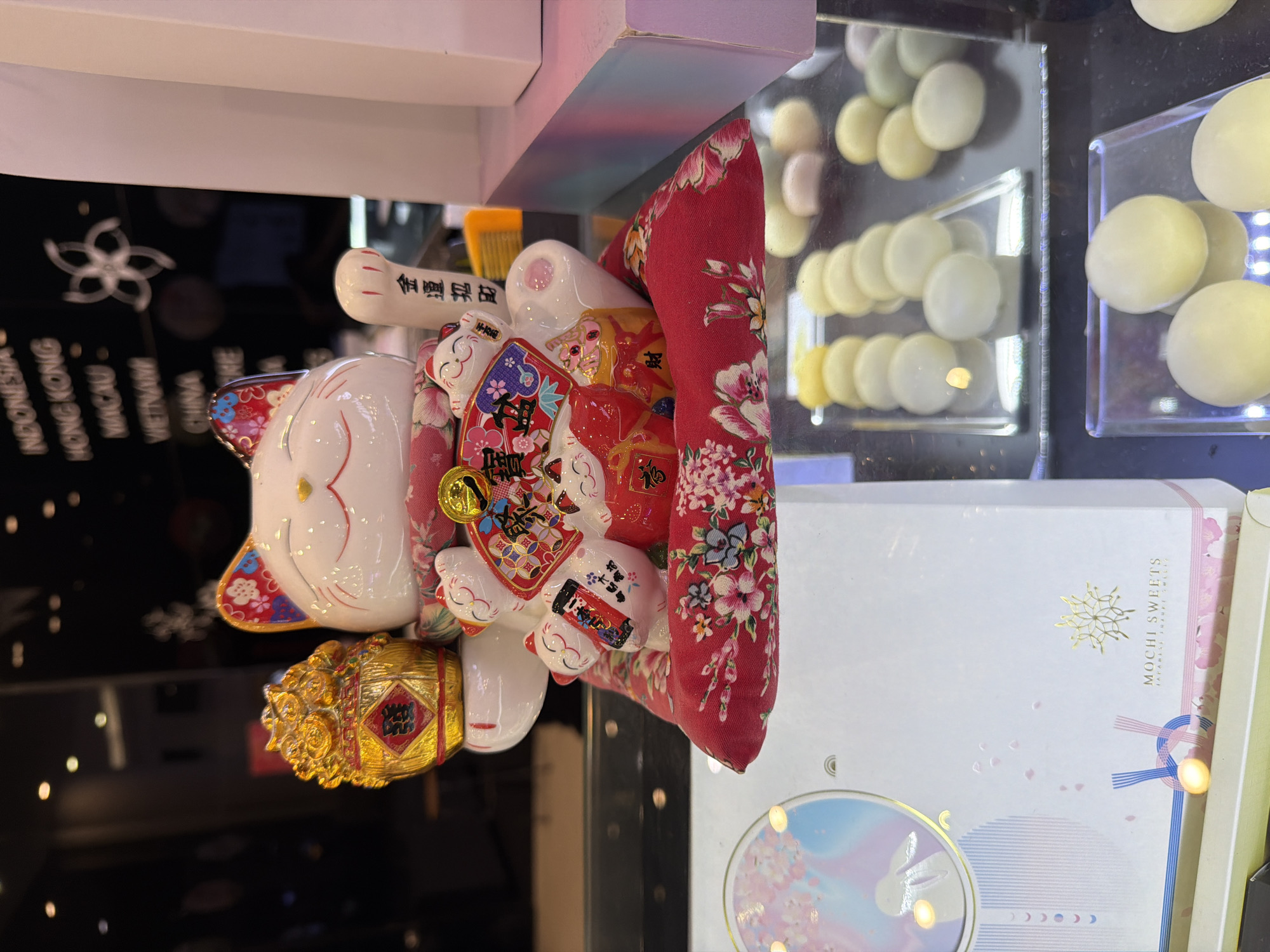 A close-up, slightly angled shot features a white Maneki-neko (beckoning cat) figurine, a popular symbol of luck in Japan and East Asia. The figurine is adorned with floral patterns on its ears and a red cushion beneath it, also decorated with a floral motif. The cat holds various auspicious items in its paws, including a golden treasure pot and a fan inscribed with characters. Behind the figurine, a blurred background shows reflections of lights and text on a dark surface, with words like "INDONESIA," "HONG KONG," "MACAU," and "VIETNAM" faintly visible, suggesting a retail or display environment. In the foreground, a white box with delicate pink and blue accents and the text "MOCHI SWEETS" is visible, along with a display case filled with round, pale-colored confections, likely mochi. The overall scene appears to be set within a sweets shop in Đà Nẵng, Vietnam, specifically in a location that might be in or near a shopping mall or a tourist-oriented area given the prominent display of international destinations in the background text. There are no people or animals directly depicted in the image. The lighting is bright, highlighting the figurine and the sweets.