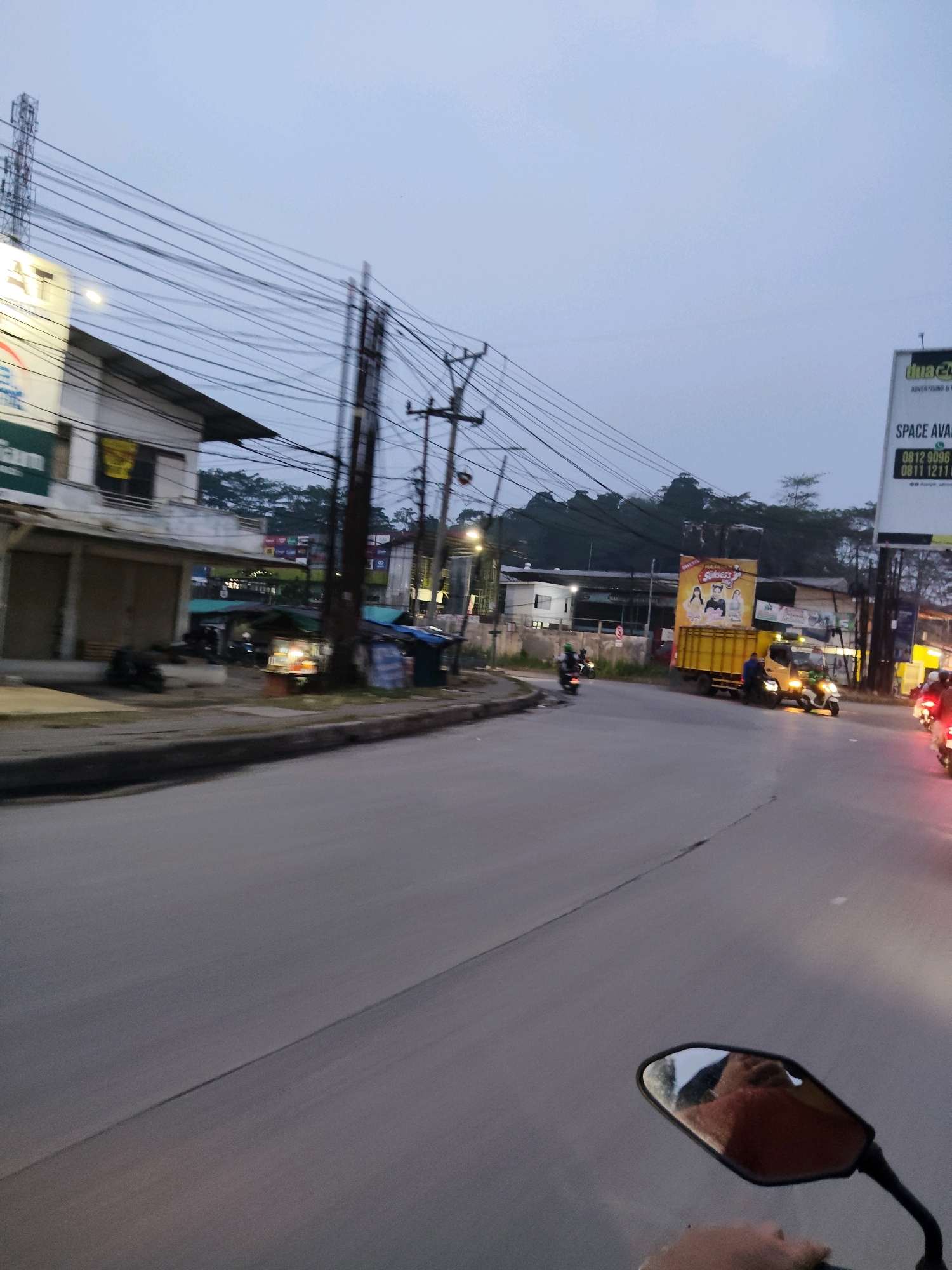 The image depicts a roadside scene in Kaduagung Tengah, Indonesia, likely taken at dusk. A paved road with a visible lane divider occupies the foreground, curving to the right. A motorcycle, from which the photo is taken, is partially visible in the bottom right corner, including its handlebar and side mirror reflecting the rider's face and torso.
Along the left side of the road, a multi-story building with closed storefronts is visible. Above the building, numerous electrical wires are strung between poles, creating a dense network overhead. A tall telecommunications tower stands to the far left.
Further down the road, a bright yellow truck with an advertisement banner on its back is parked. A person in dark clothing is standing next to the truck. Several other vehicles, including motorcycles with illuminated taillights, are on the road, suggesting ongoing traffic. Small stalls or shops with awnings are present along the sidewalk. The background shows a line of trees and hills under a pale, overcast sky. The overall impression is that of a busy, developing urban or suburban environment transitioning into evening.