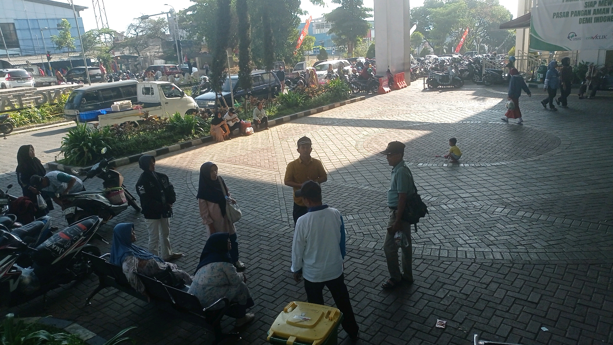 The image depicts a street scene in Batu, Indonesia, likely near a market or public building. In the foreground, several people are gathered, some standing and others seated on a bench. One woman wearing a blue hijab is sitting on the bench, facing right. Another woman in a patterned hijab is also seated. A man in a yellow shirt and a cap is standing and speaking to others. To the left, there are several motorcycles parked, some with people attending to them or standing nearby. A man in a green shirt and a cap is walking away from the camera on the right side of the image. Further back, a young child in a yellow shirt is playing on the ground. The area is paved with interlocking bricks, and there are trees and buildings in the background, suggesting an urban environment. A large banner with text is visible on a building in the distance. The lighting suggests it is daytime, with shadows cast on the ground.