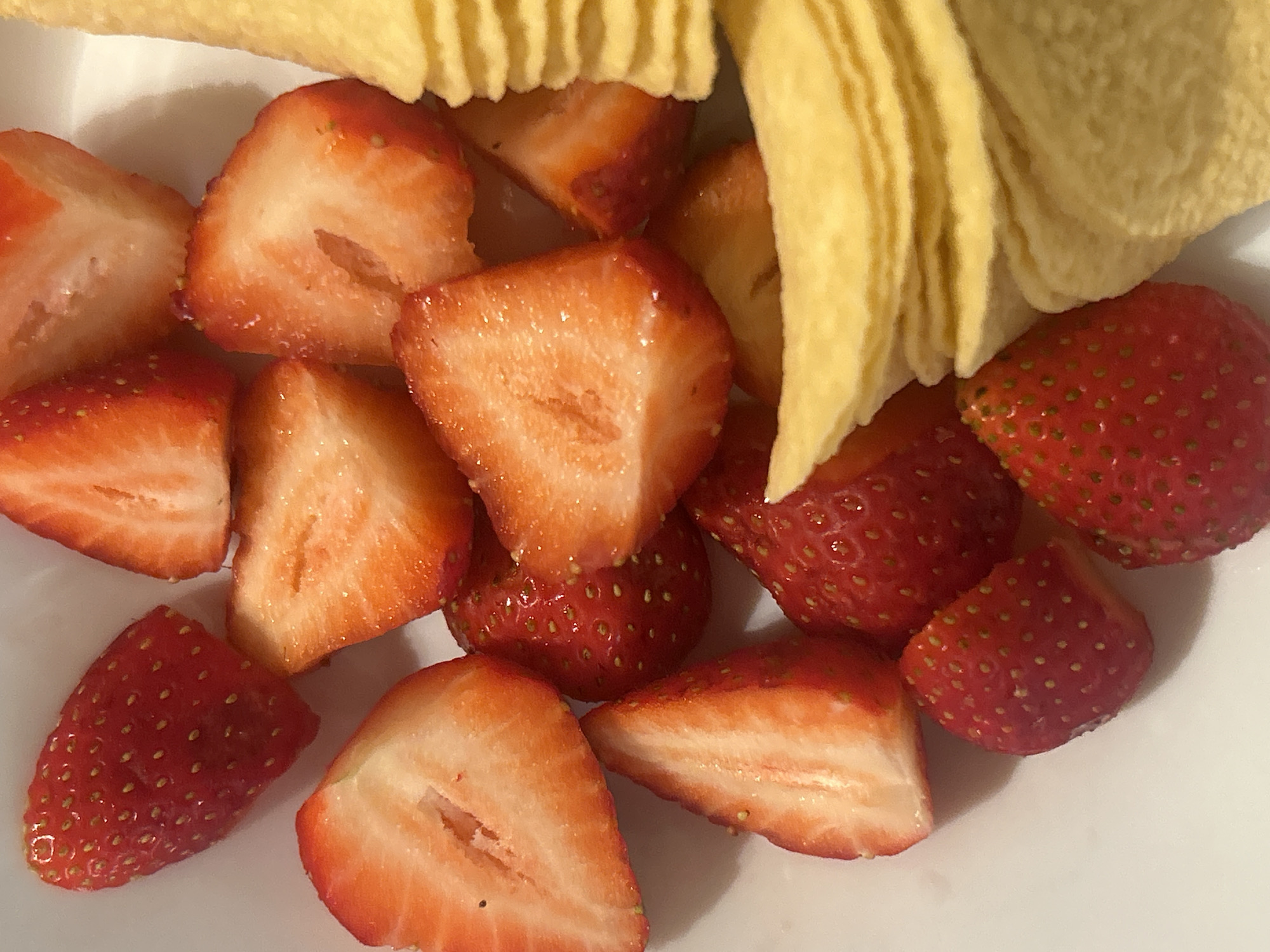 A close-up shot shows a white plate containing cut strawberries and potato chips. The strawberries are bright red and have been sliced into various shapes and sizes. The potato chips are yellow and arranged in a cluster. The background is out of focus.